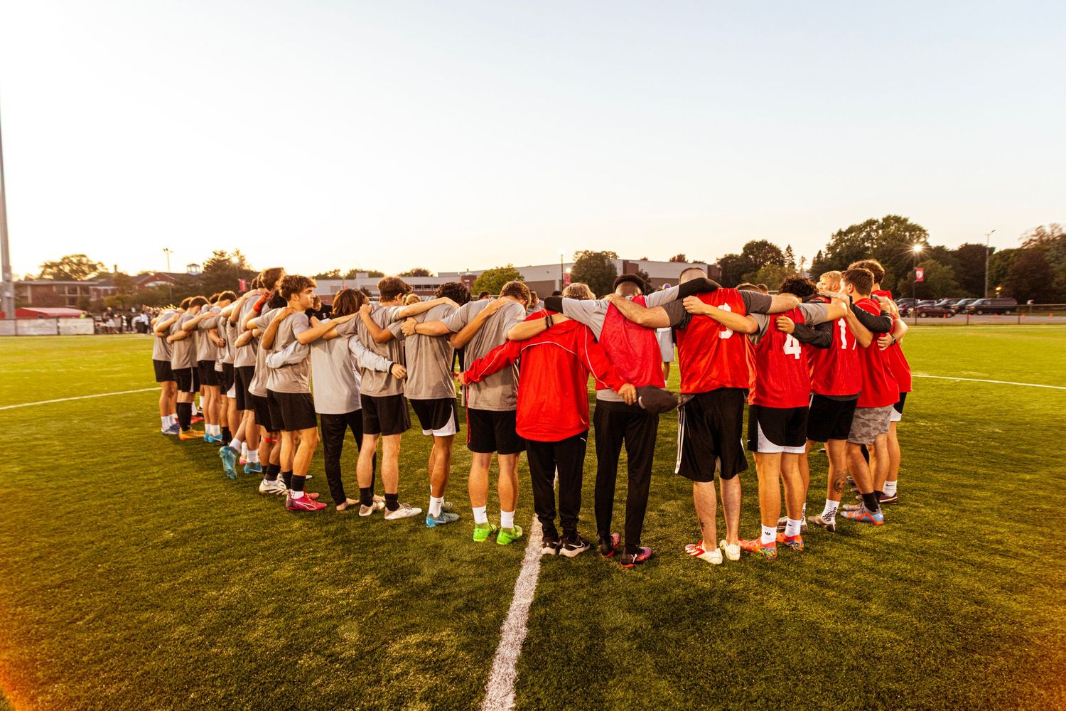 Sports team huddled in a circle on a grass field at sunset with coaches in red shirts.