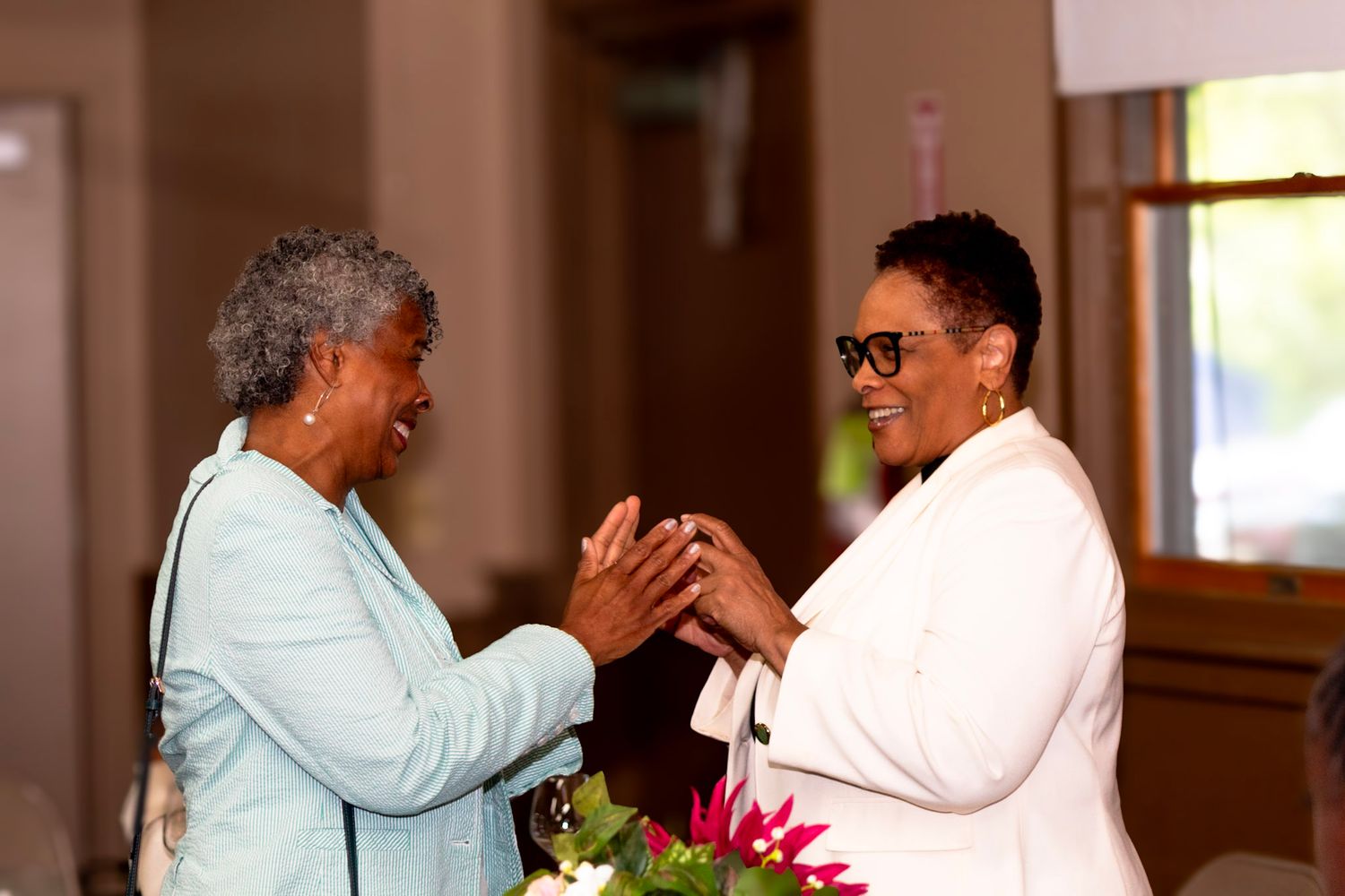 Two women in light colored outfits share a joyful moment together with animated hand gestures.