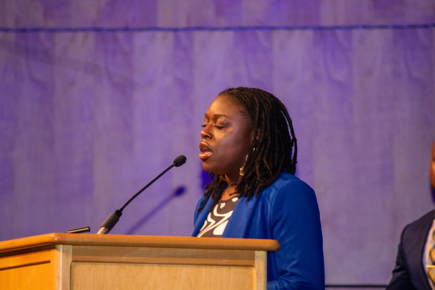 Speaker at podium wearing bright blue blazer against purple backdrop.