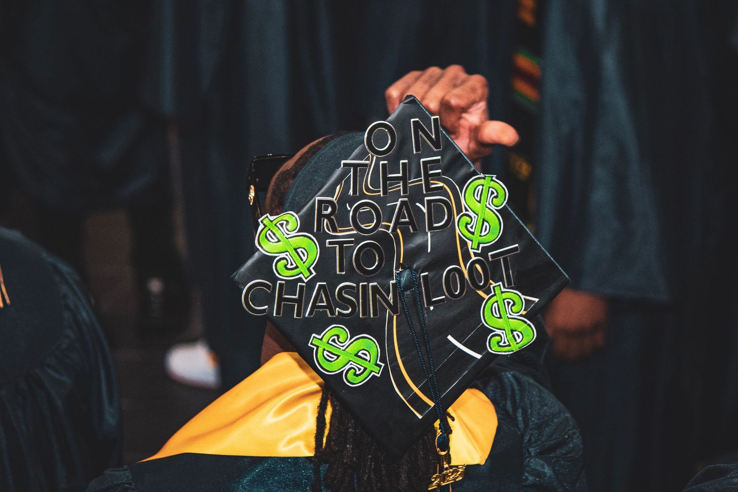 Decorated graduation cap with green money-themed design against dark background.