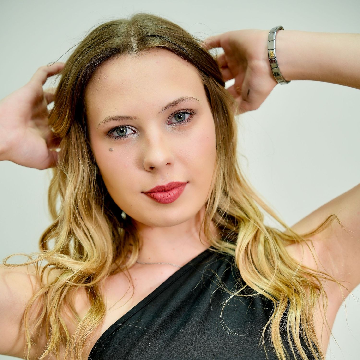 A woman with ombre blonde hair poses in a black top while running fingers through her wavy locks.