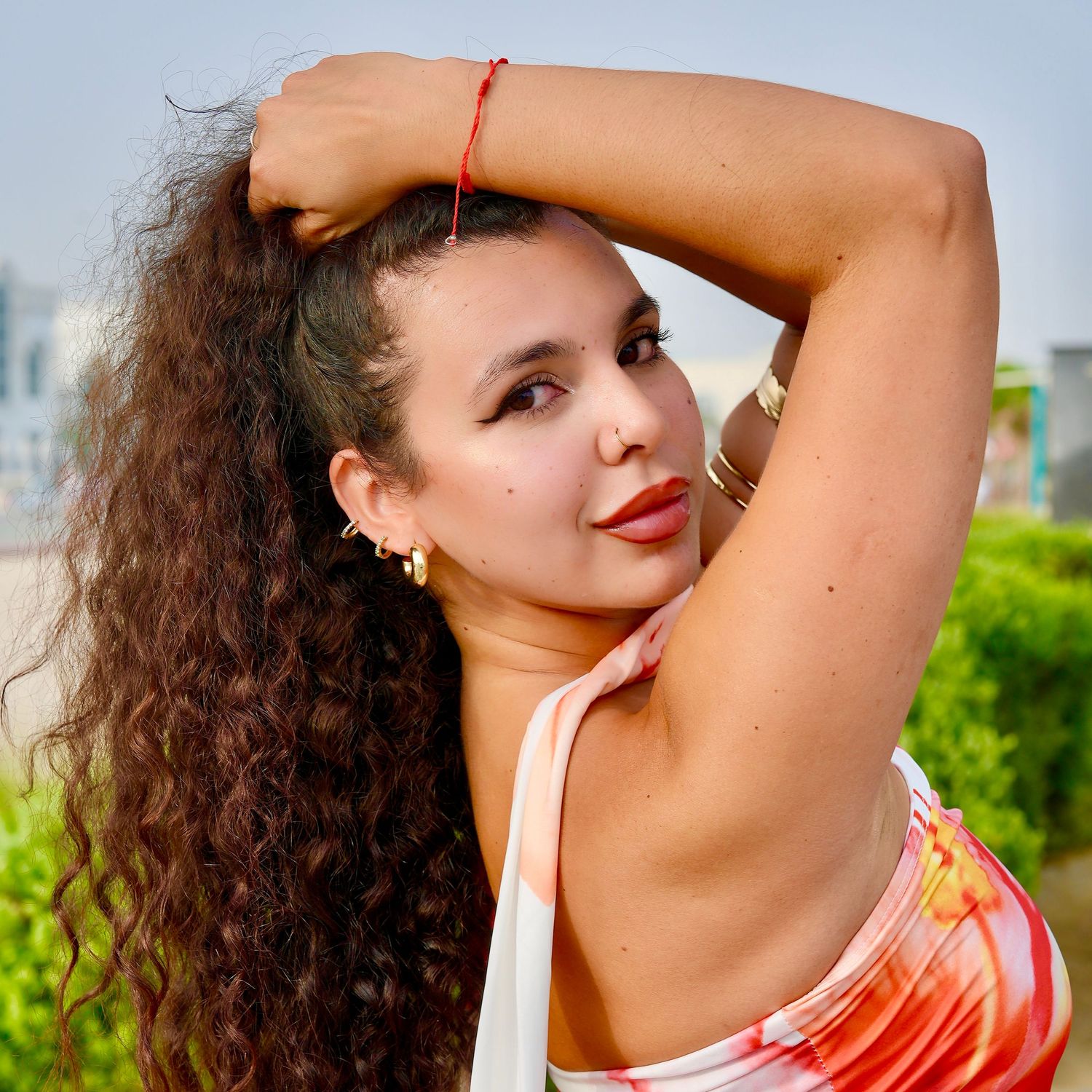 Person with curly hair wearing a coral-colored top poses outdoors against a bright sky and greenery.