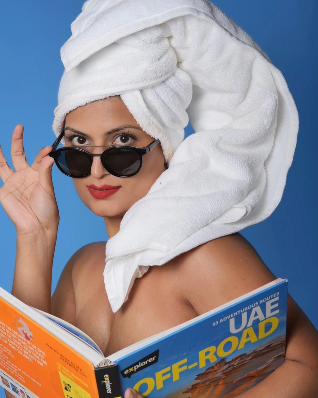 A glamorous figure in sunglasses and white towel turban reads an off-road UAE guidebook against a blue background.