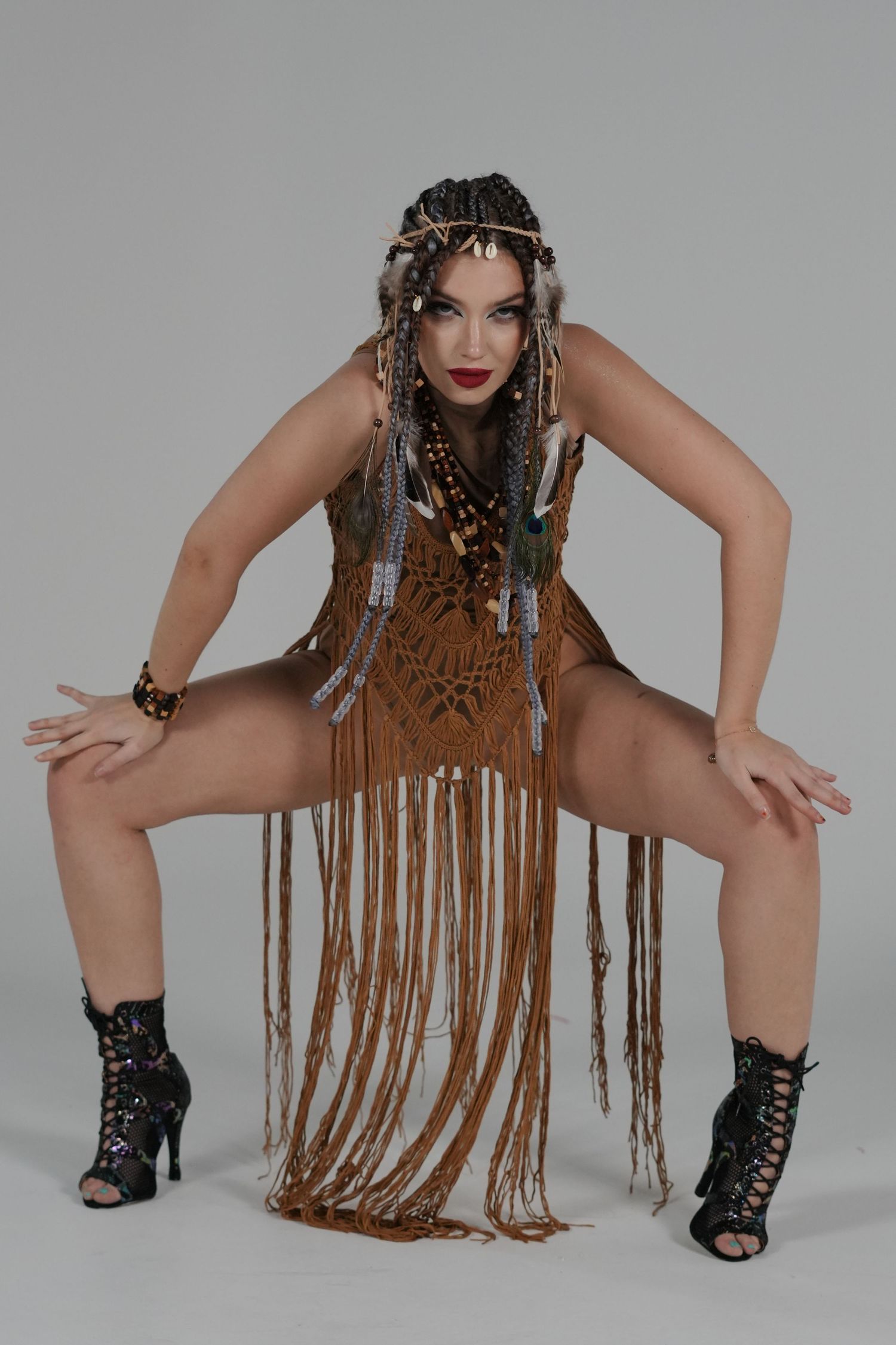 Model in tribal-inspired fringe outfit and black boots poses dramatically against gray backdrop in fashion photo series.