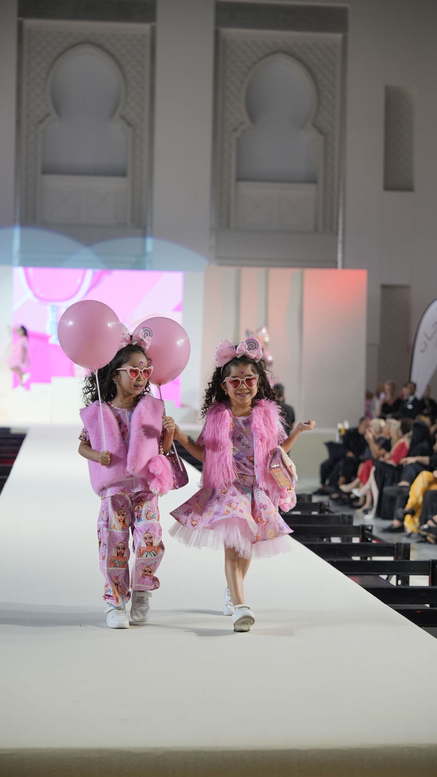 Young models walking runway in pink outfits at fashion show.