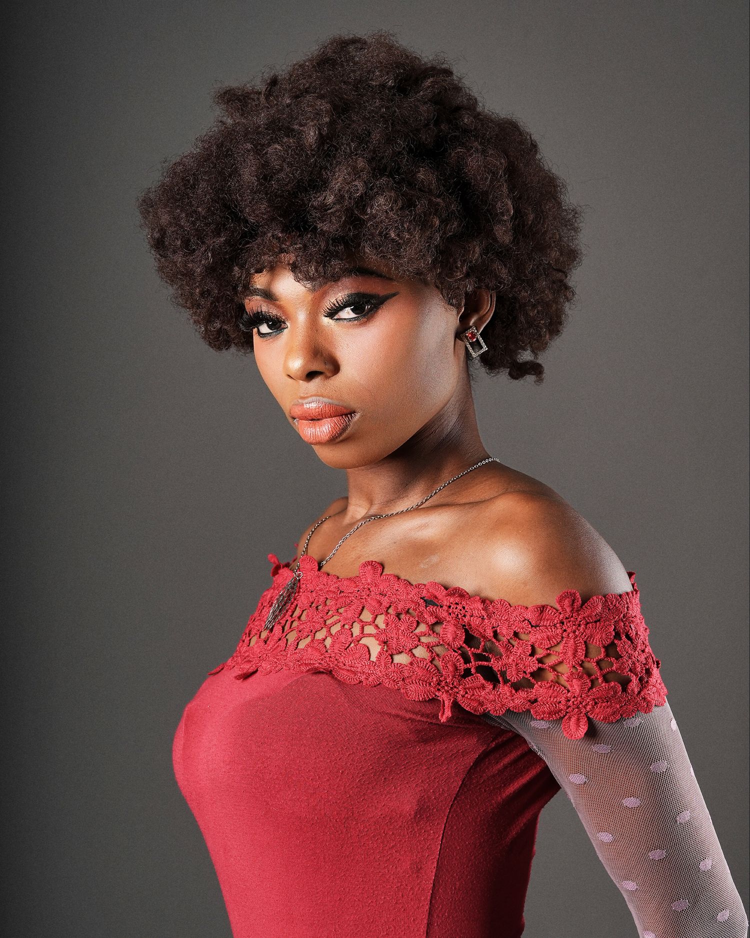 Young woman with natural afro hair wearing a red off-shoulder crochet top, posing confidently against a gray background.