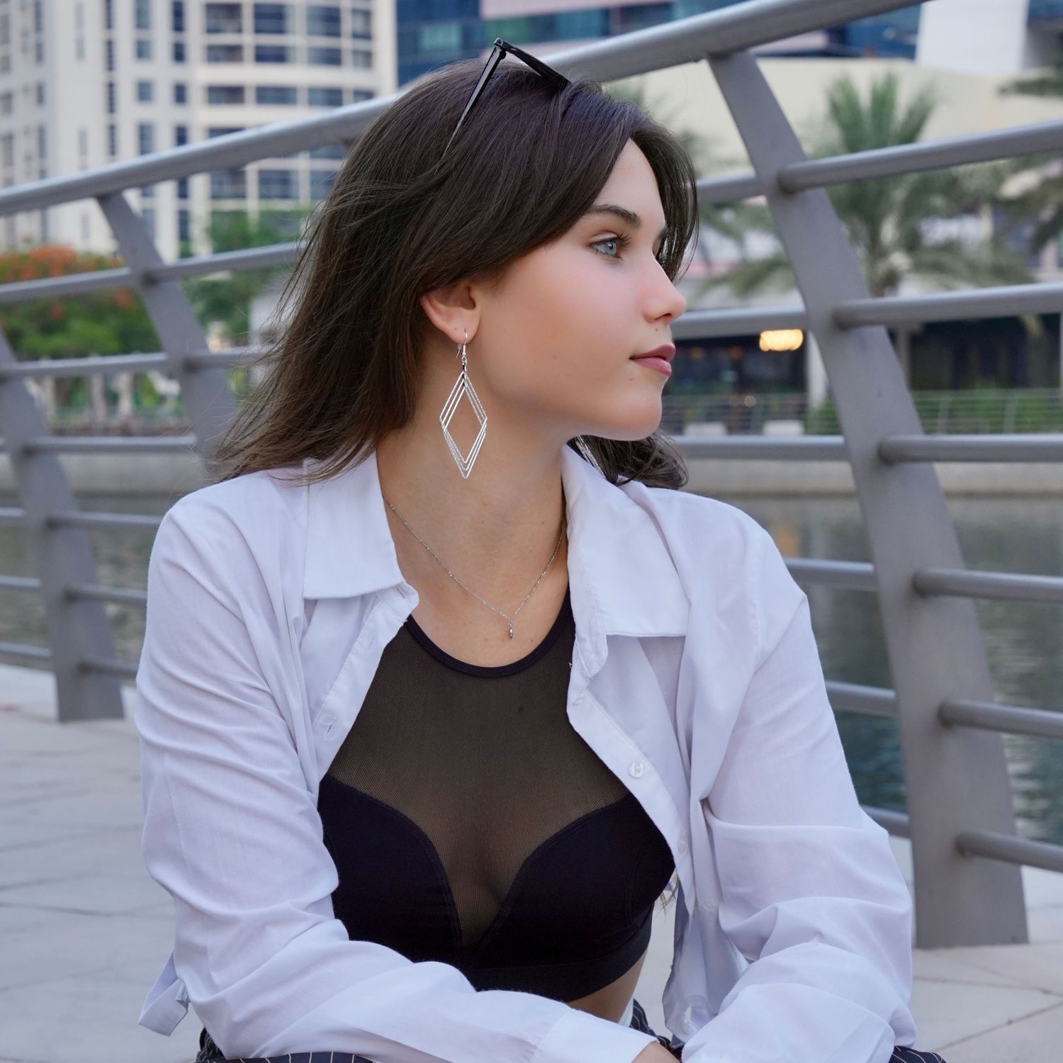 Woman in white blazer and black top poses outdoors against modern urban backdrop with palm trees.