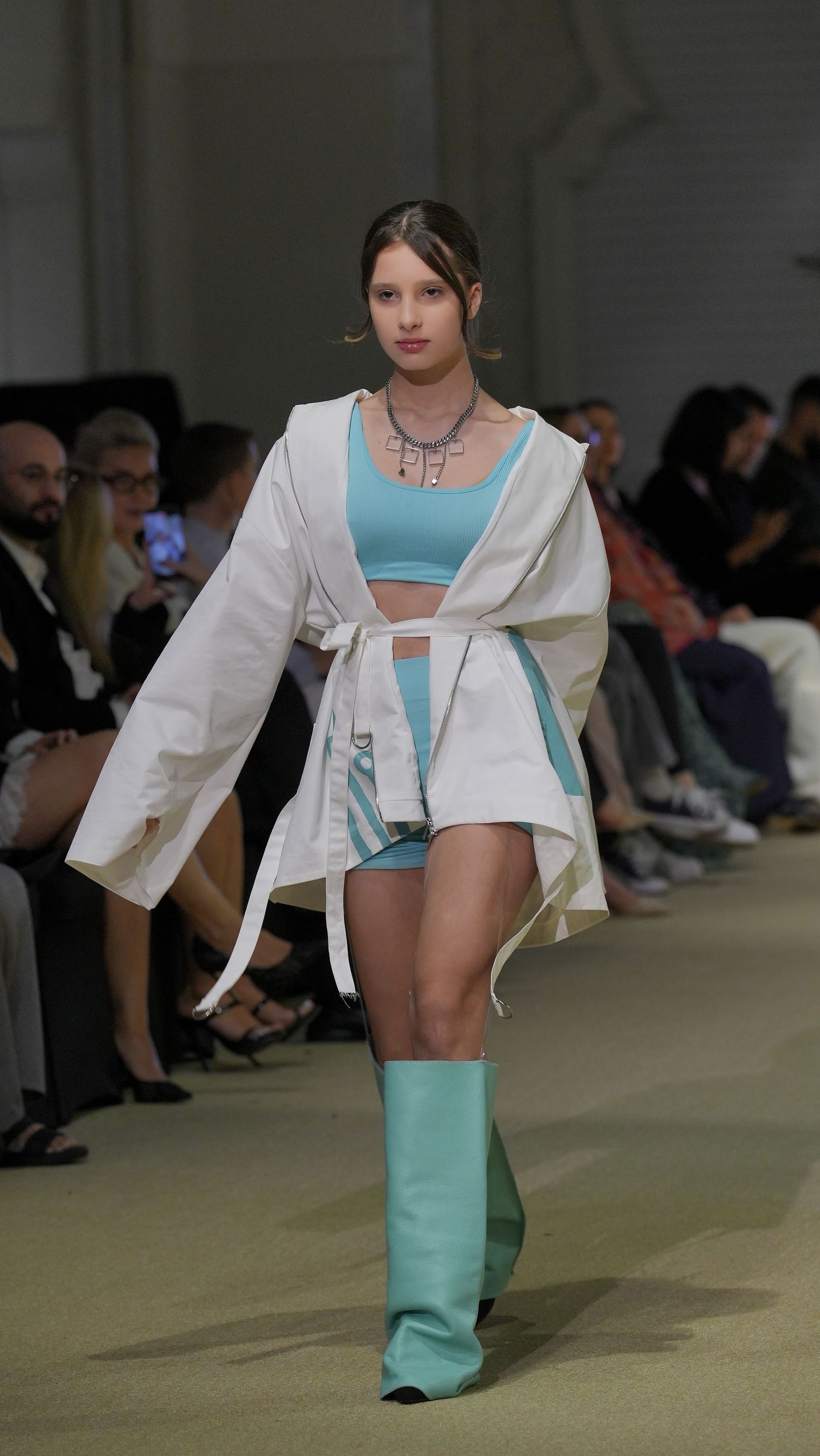 A fashion runway model walks in a white jacket and turquoise pants during a fashion show presentation.