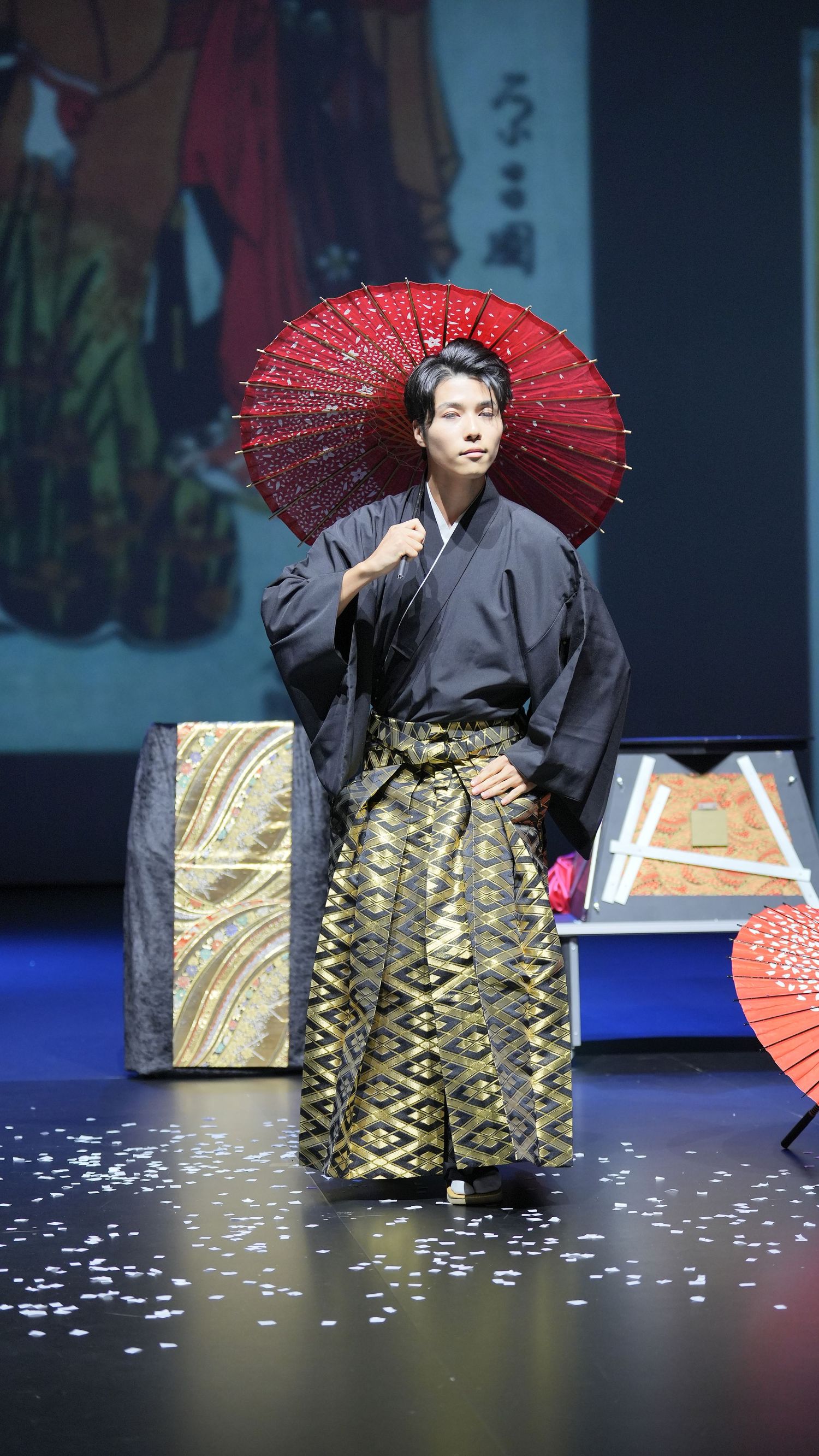 Traditional Japanese performer in dark kimono and hakama holds a red paper umbrella on stage with scattered petals.