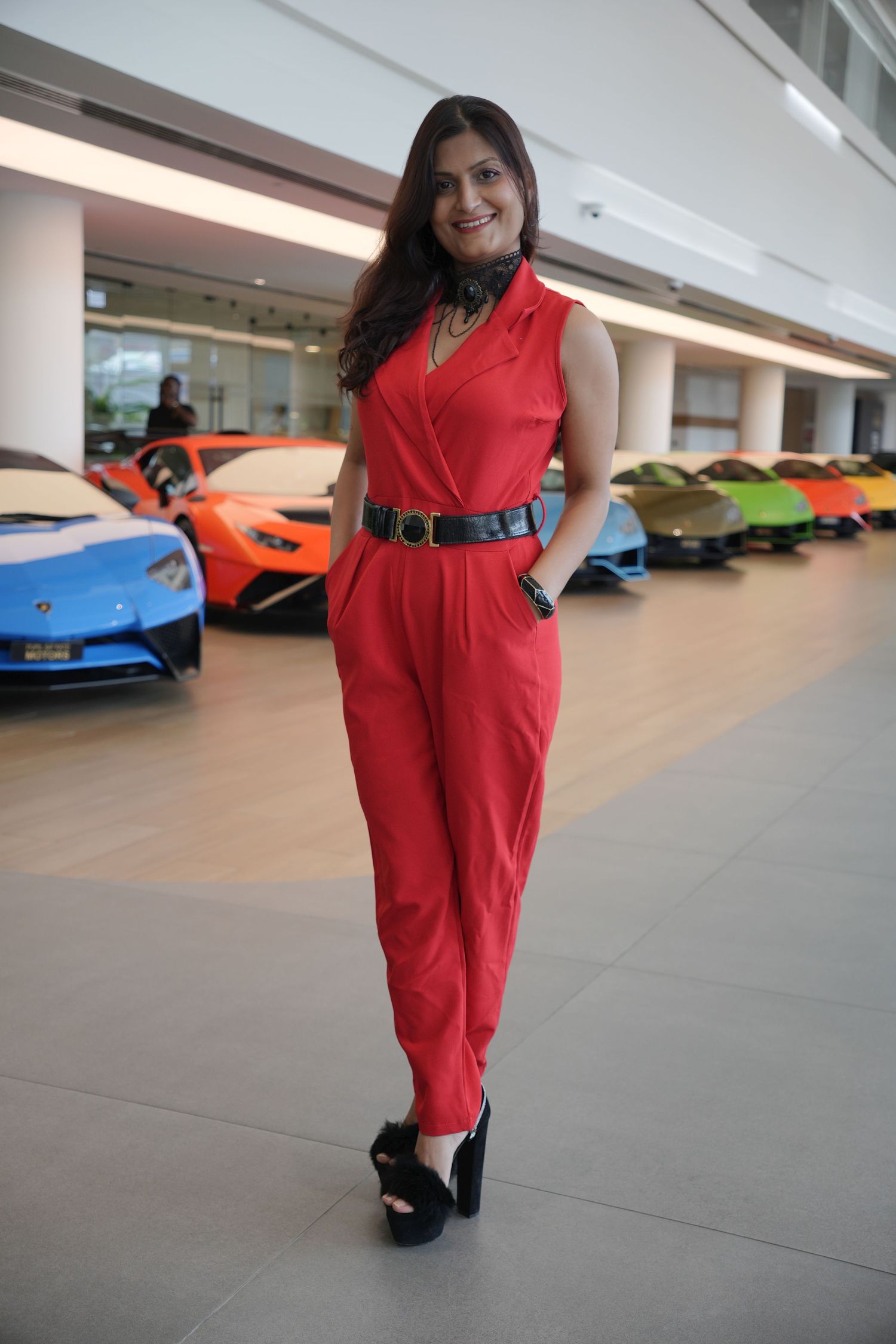 Person in a sleeveless red jumpsuit poses in front of luxury cars in a showroom.