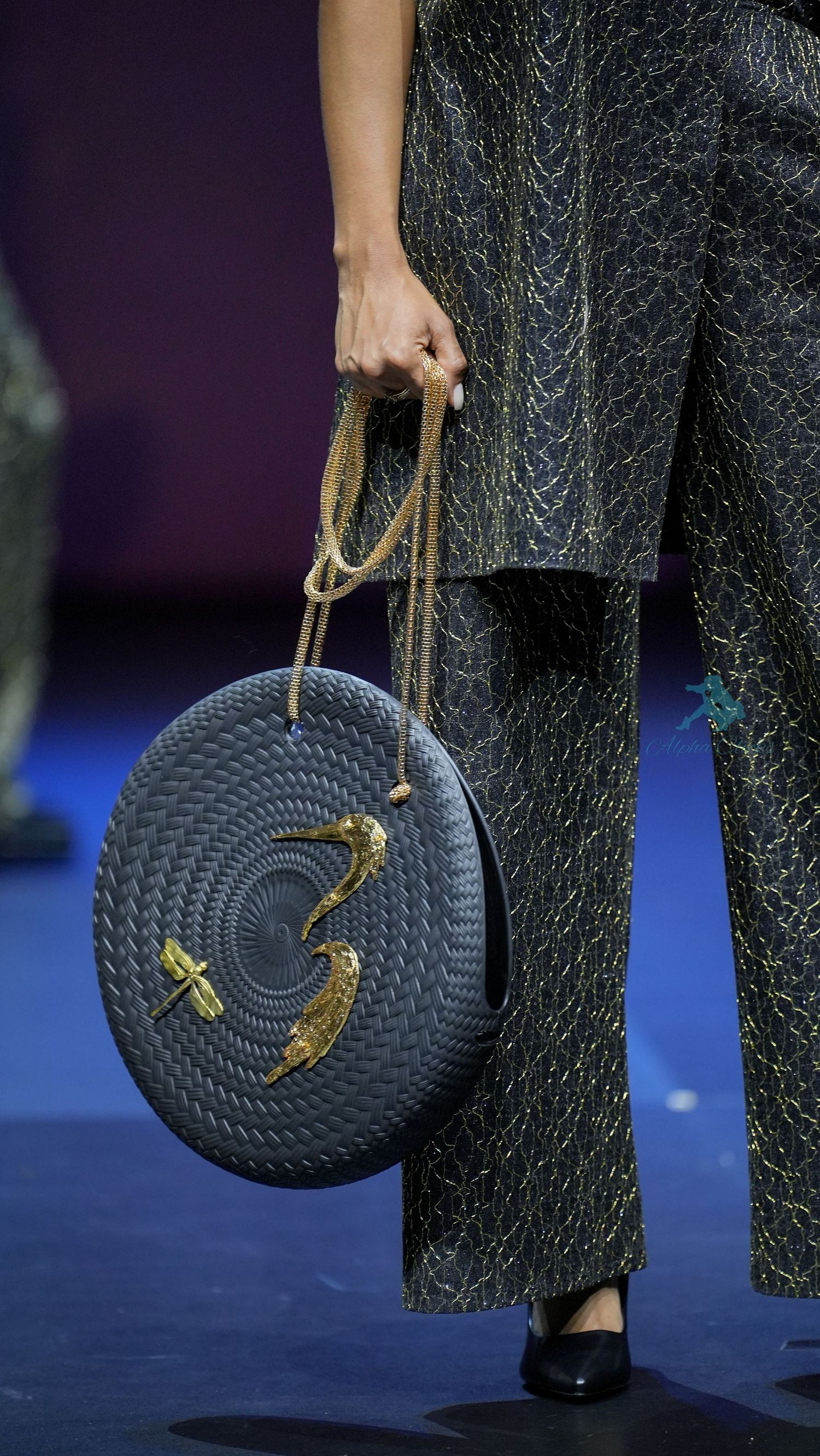 A black woven circular handbag with gold hardware swings against a dark textured fabric background on a runway.