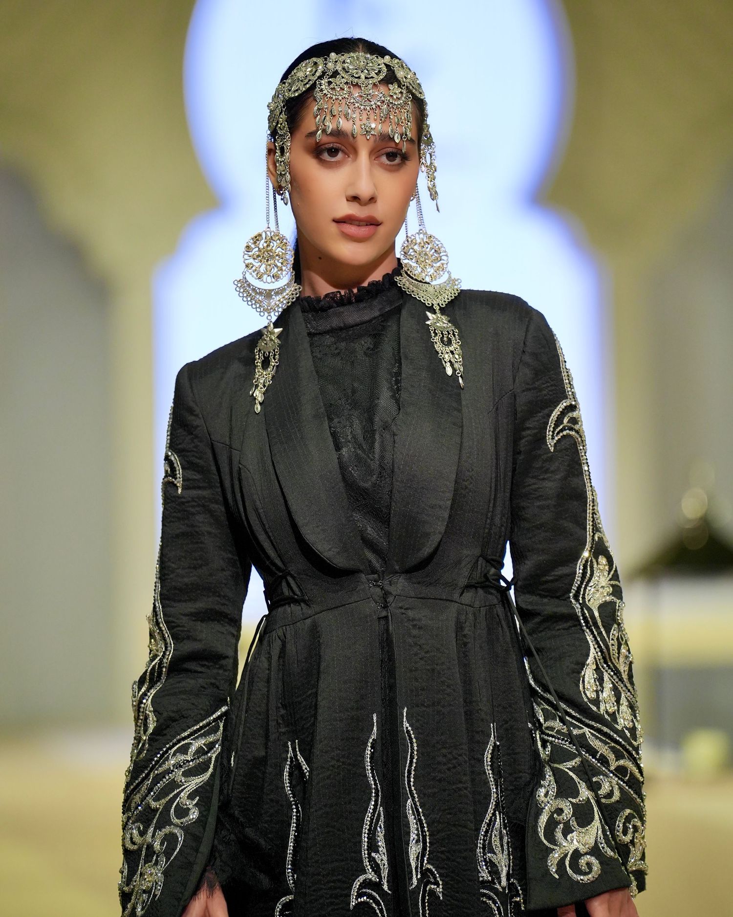 Model walks runway in black structured jacket with ornate gold embroidered sleeves during fashion show presentation.