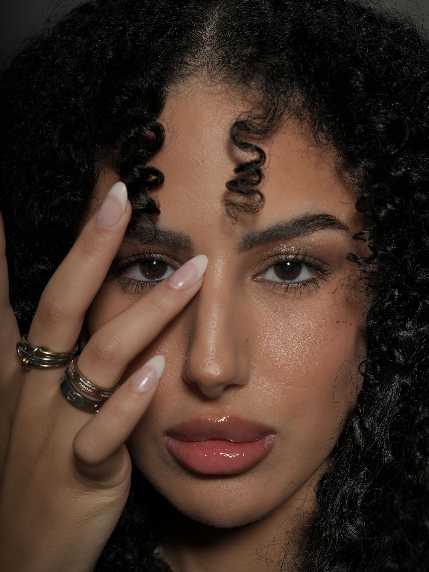 Close-up beauty portrait showing glossy lips, curly hair, and silver rings against dark background.