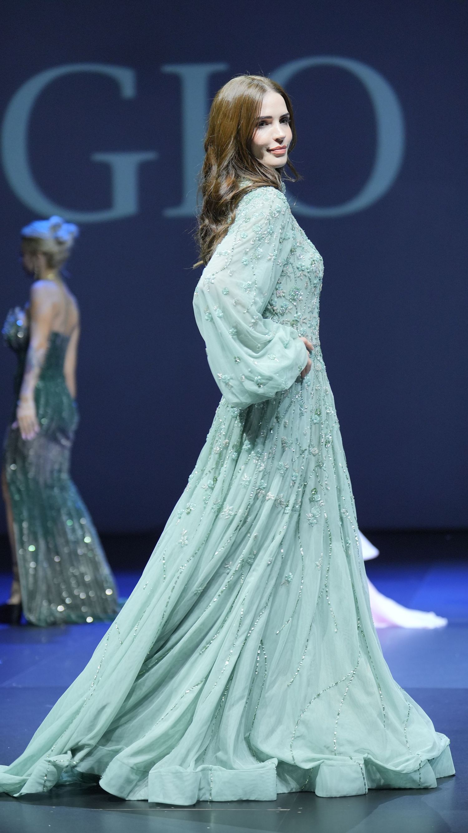 Model walks runway in flowing mint green gown with sparkly embellishments at fashion show.