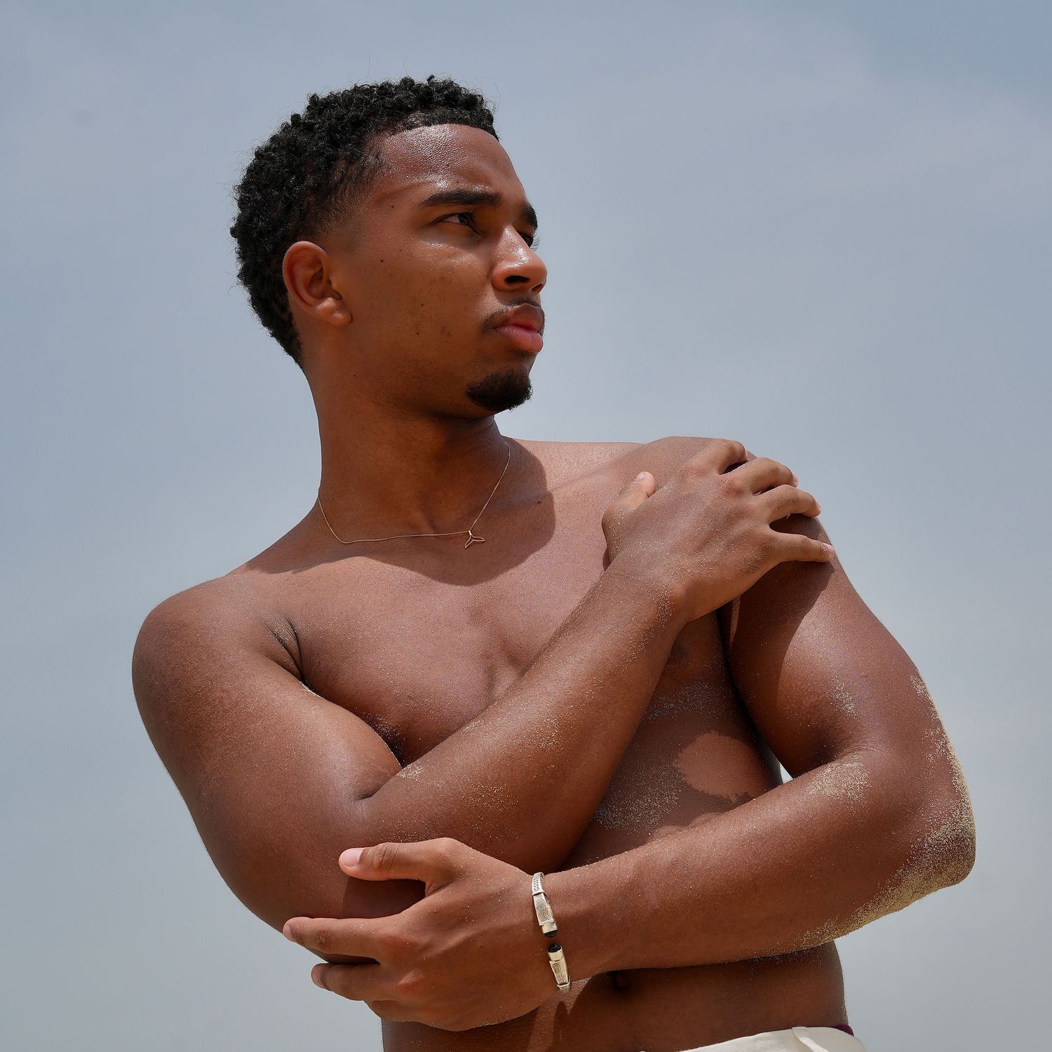 A shirtless athletic man with crossed arms poses outdoors against an overcast sky.