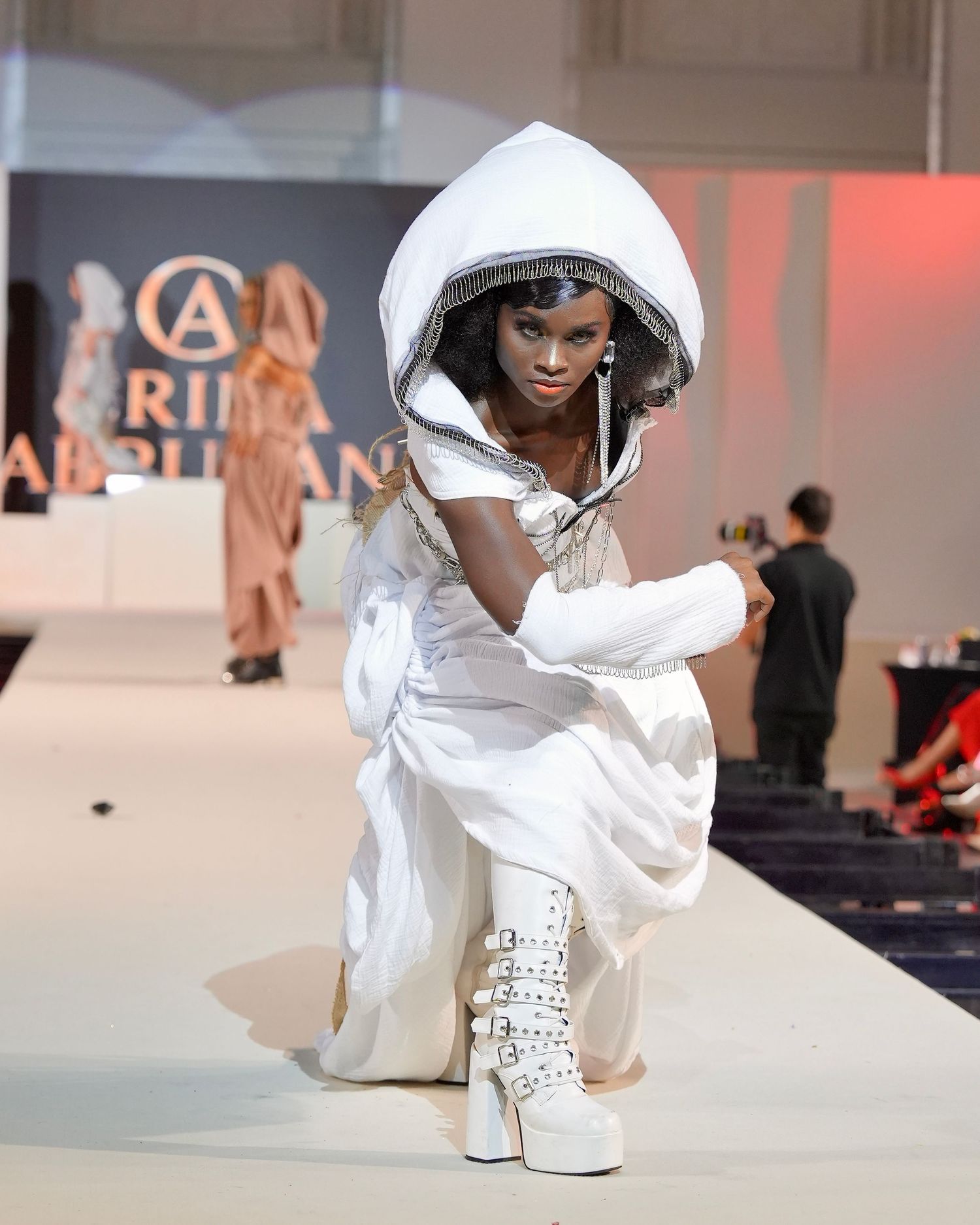 Model in white hooded ensemble with platform boots poses dramatically on fashion runway.