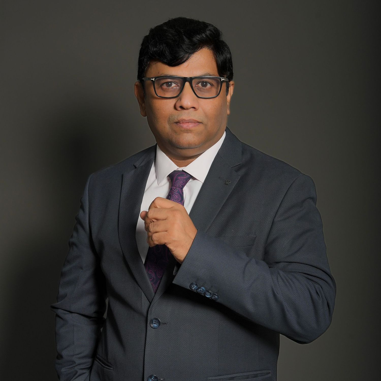 Professional headshot of someone in a dark suit and glasses against a black background in contemplative pose.