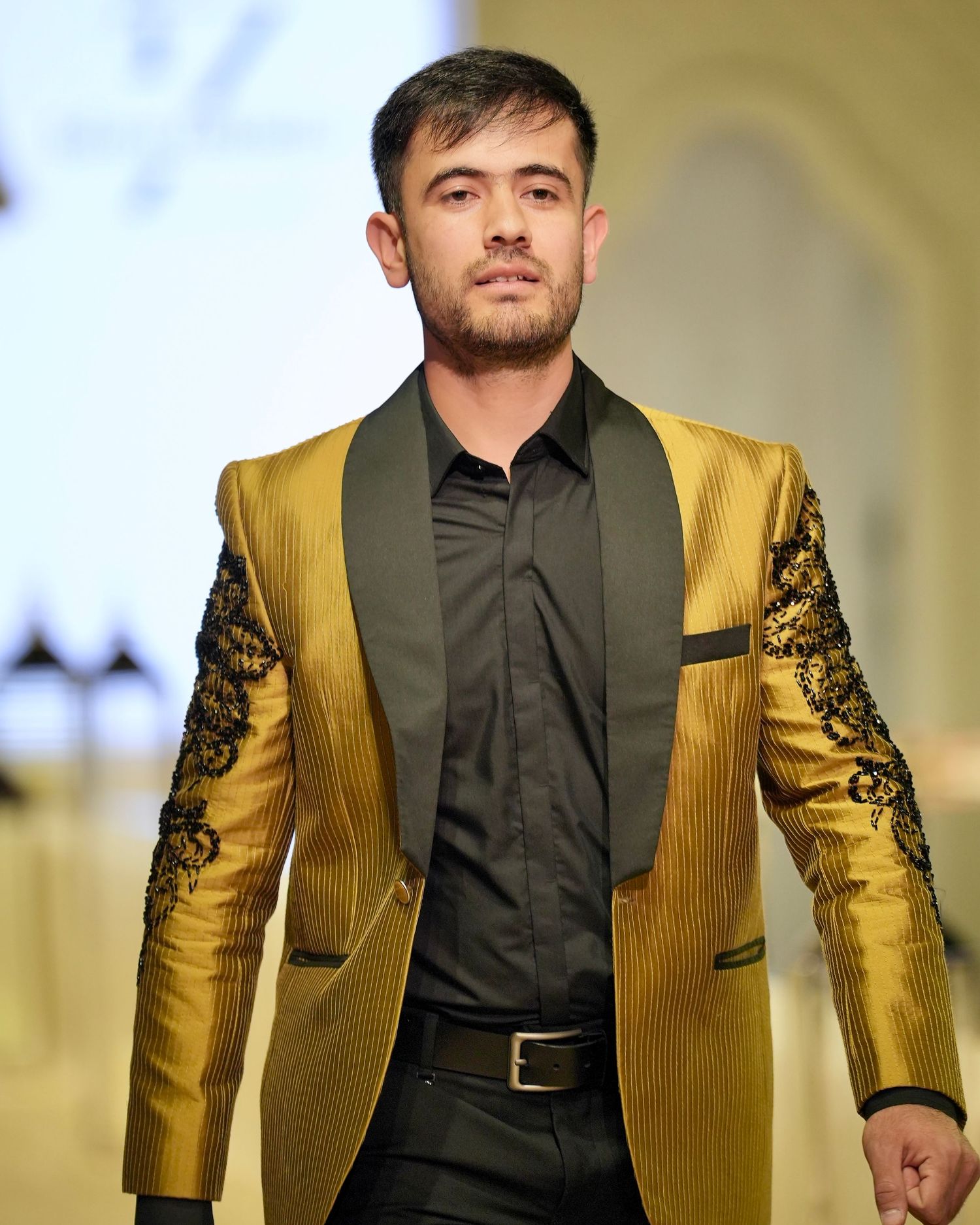 A person walks on a fashion runway wearing a metallic gold blazer with black embellishments and a black shirt.