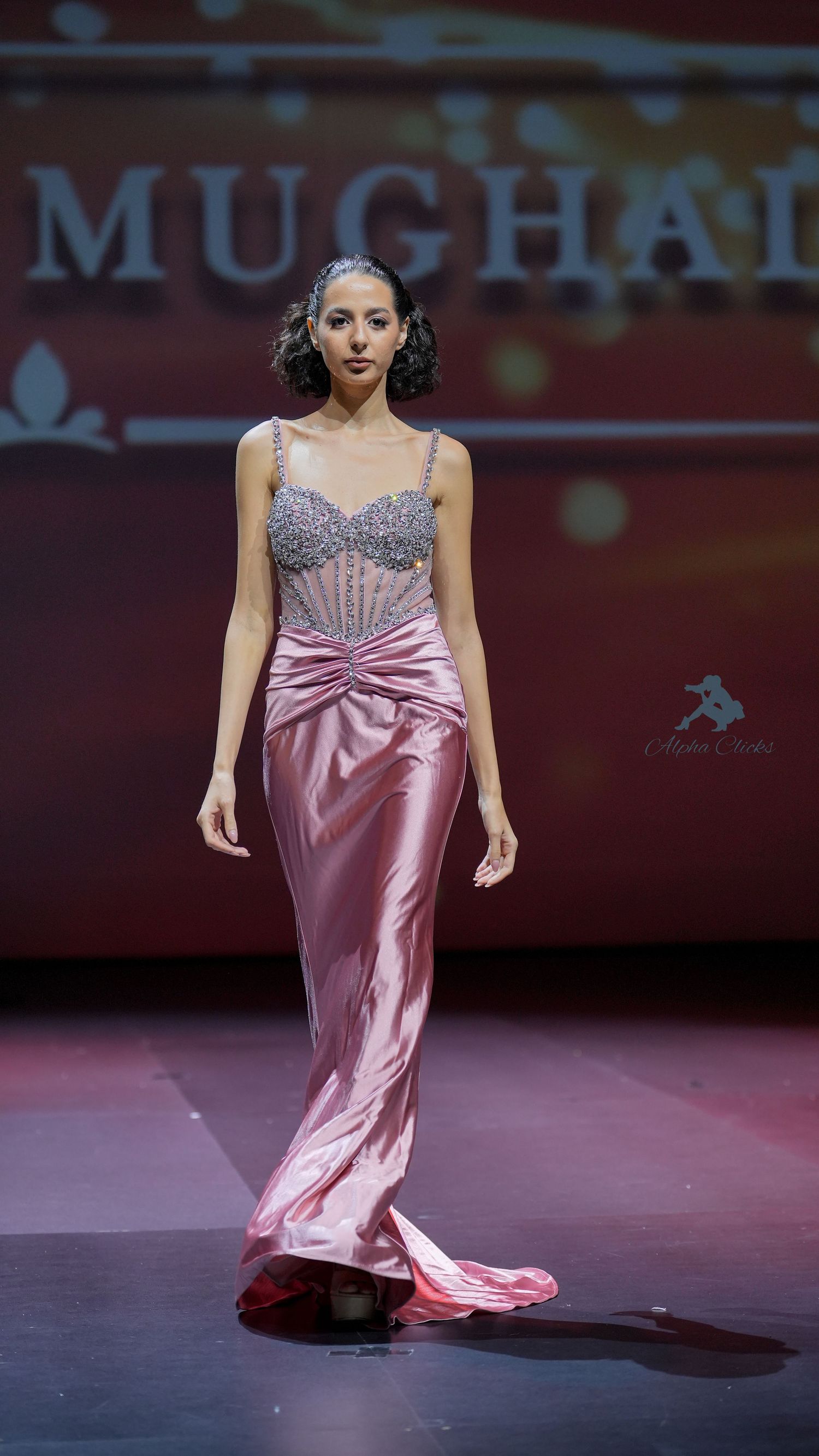 Model walks runway in flowing pink satin evening gown with crystal-embellished bodice during fashion show.
