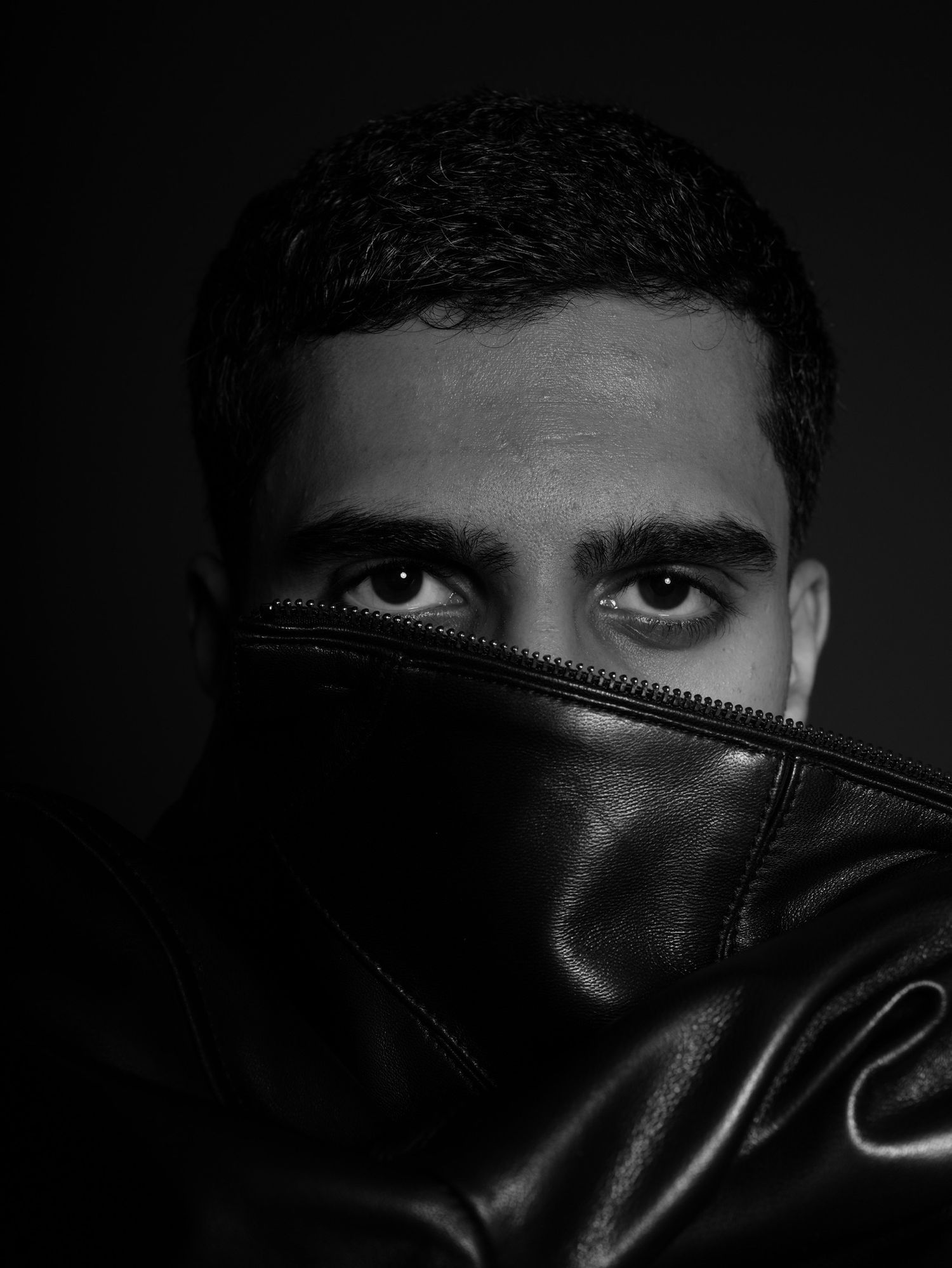 A dramatic black and white portrait showing intense eyes peering over a black leather jacket in shadow.