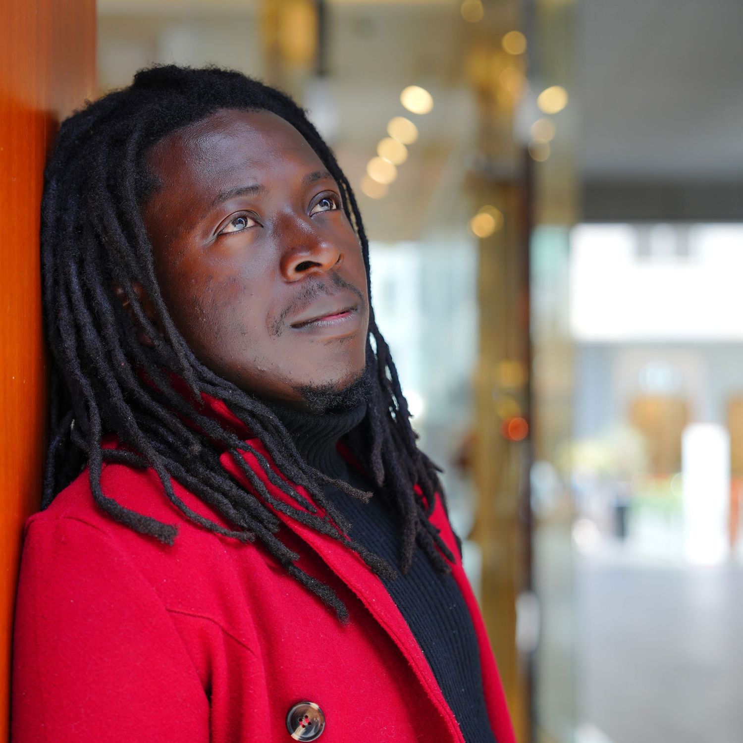Person in red jacket with dreadlocks smiling while looking up and to the side against blurred background.