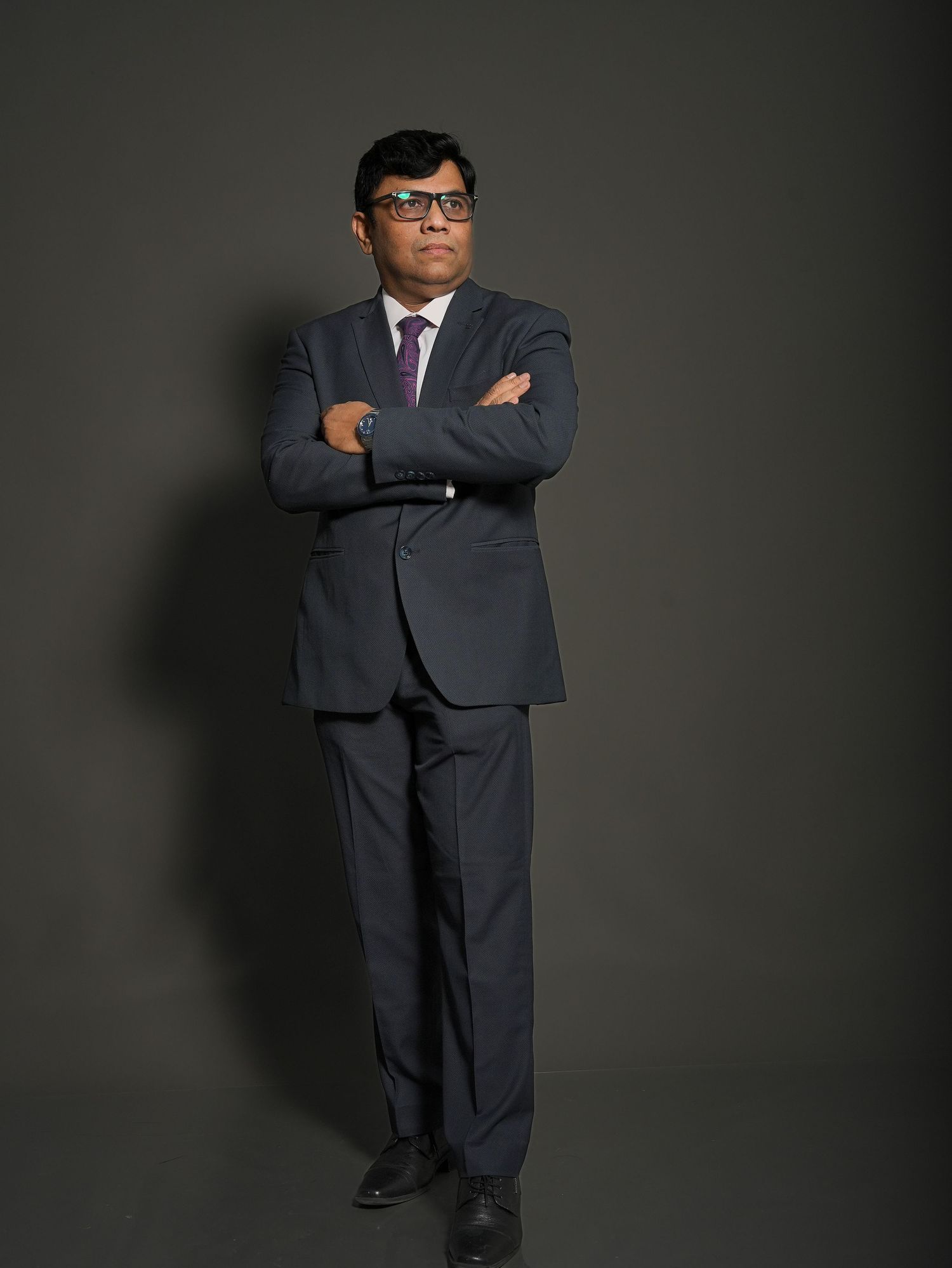 A person in a dark business suit and glasses stands with arms crossed against a shadowy gray background.