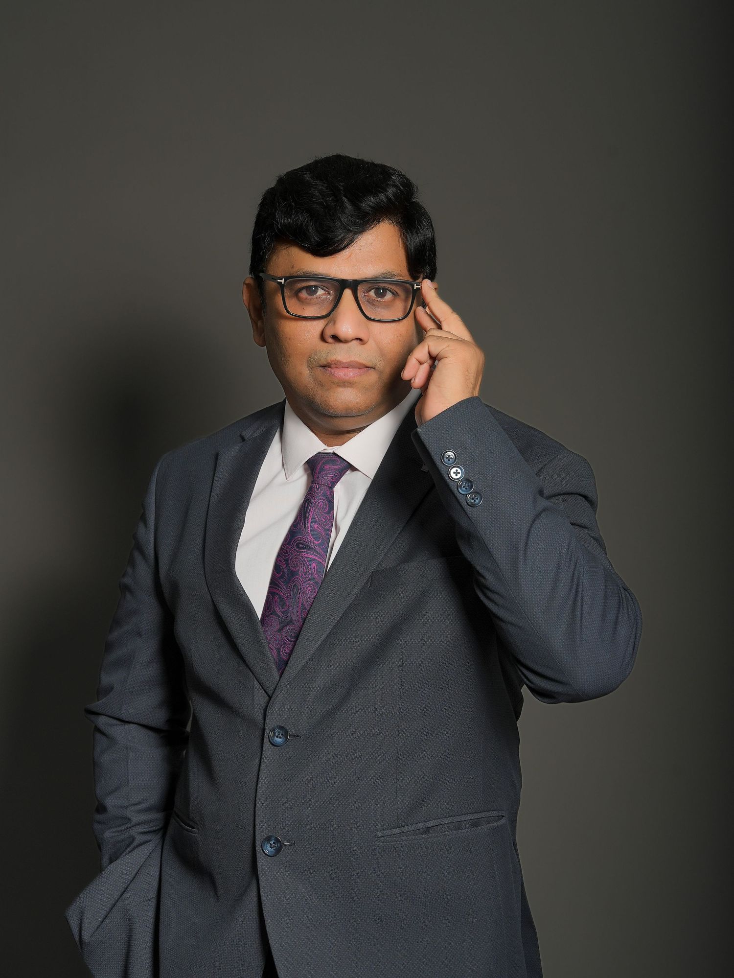 Professional man in gray suit and purple tie wearing glasses, posing confidently against a dark background.