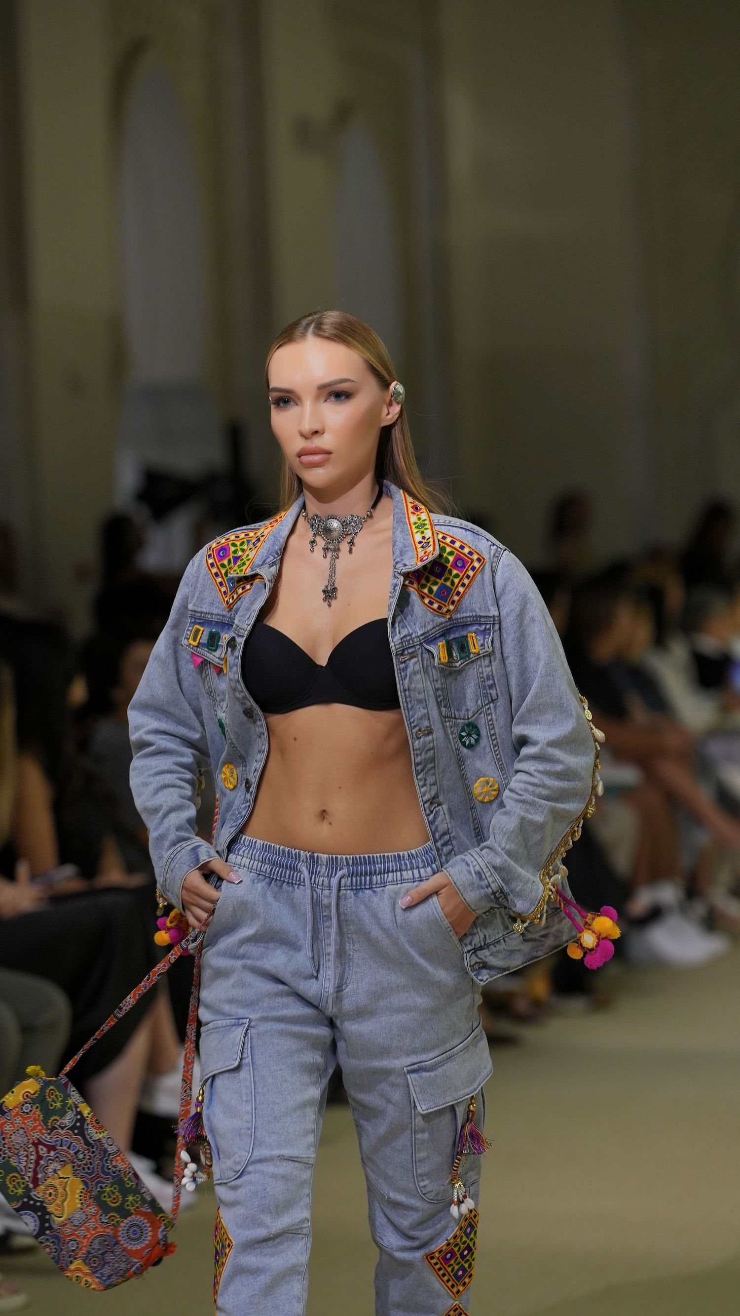 Model walks runway in light denim streetwear ensemble with colorful patches and black crop top during fashion show.