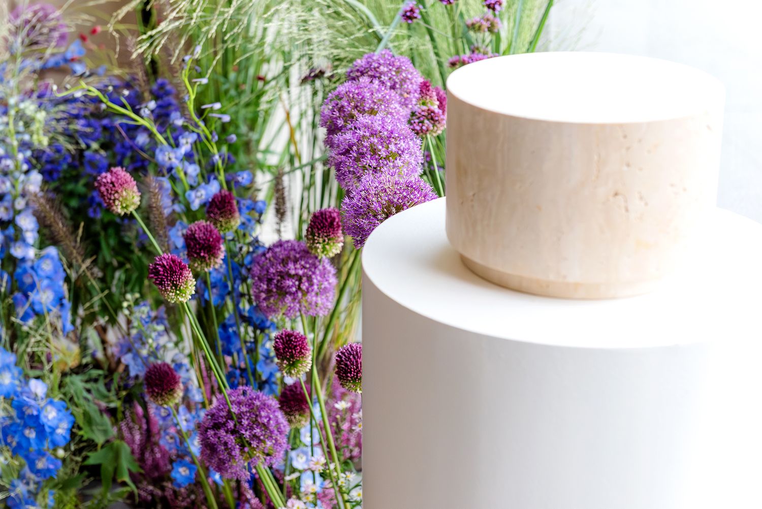 A wooden urn sits on a white pedestal surrounded by purple allium and blue delphinium flowers.