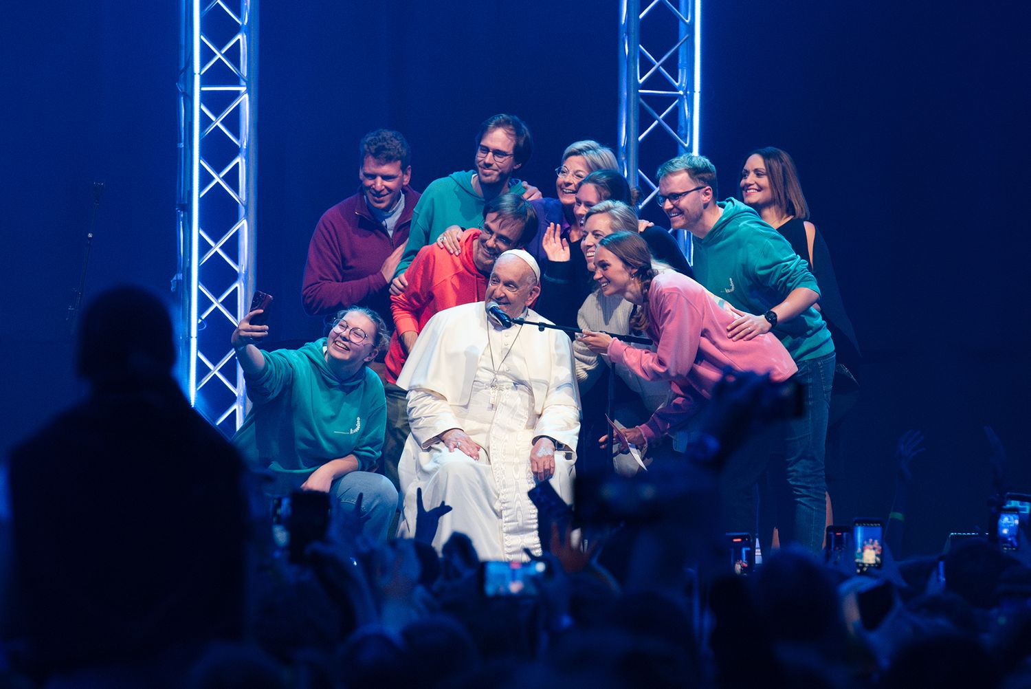 A group of people gather on a blue-lit stage around a figure dressed in white papal robes.