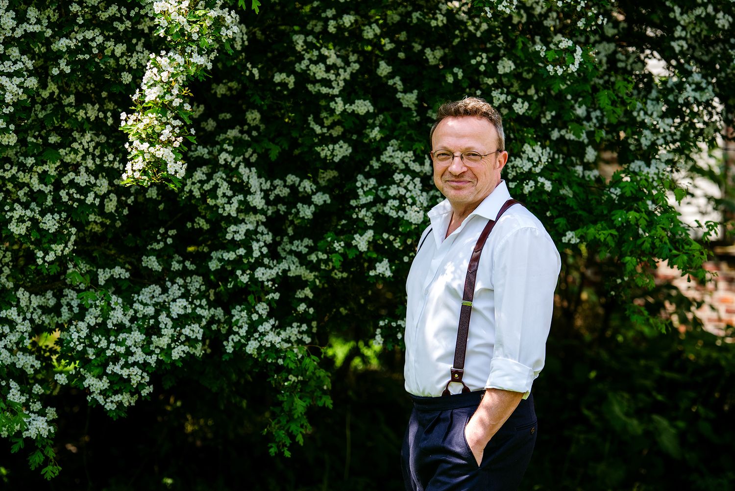 Smiling man wearing glasses and suspenders standing before blooming white flowering bushes outdoors.