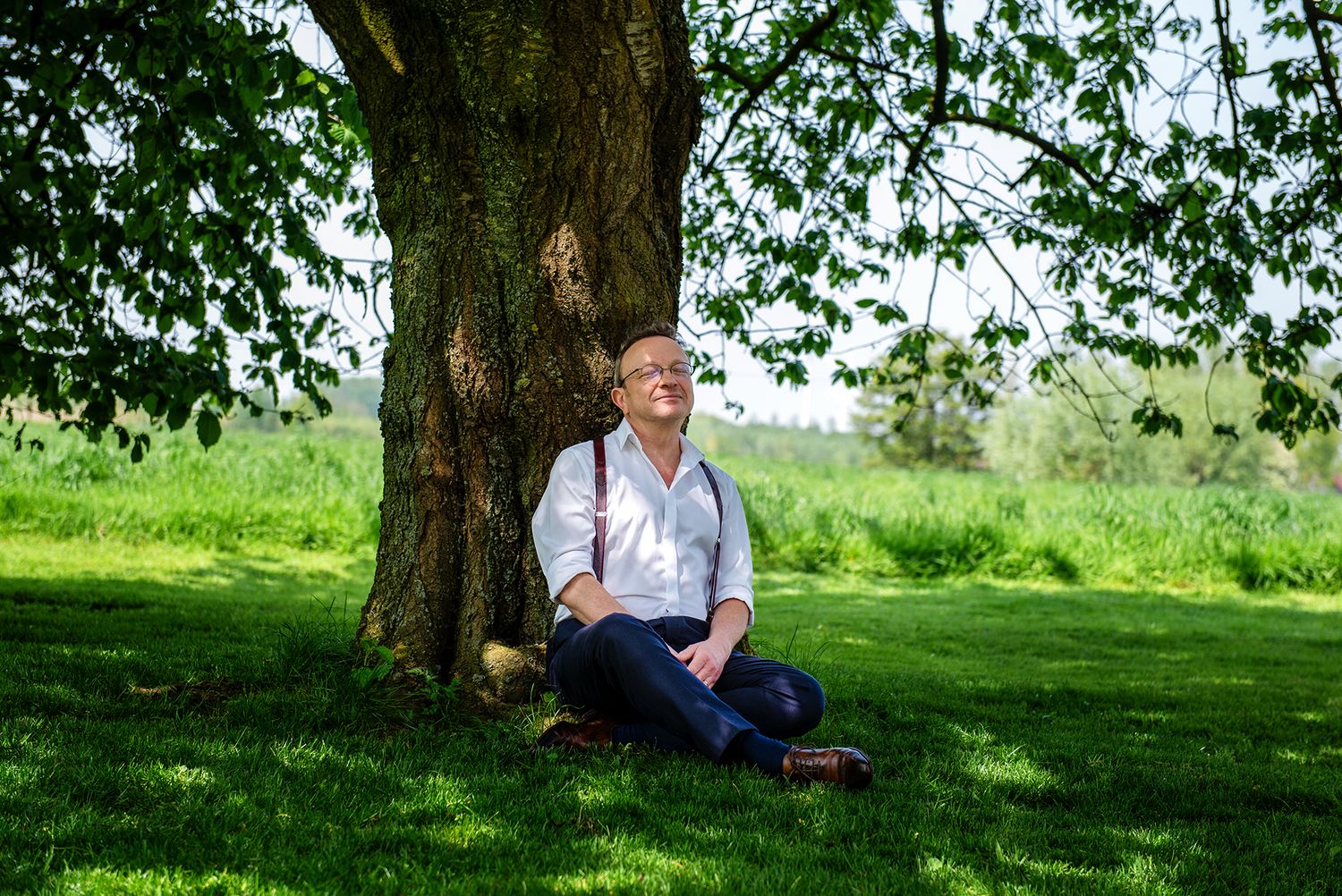 Man in white shirt and suspenders sitting relaxed against a large tree in a lush green meadow.