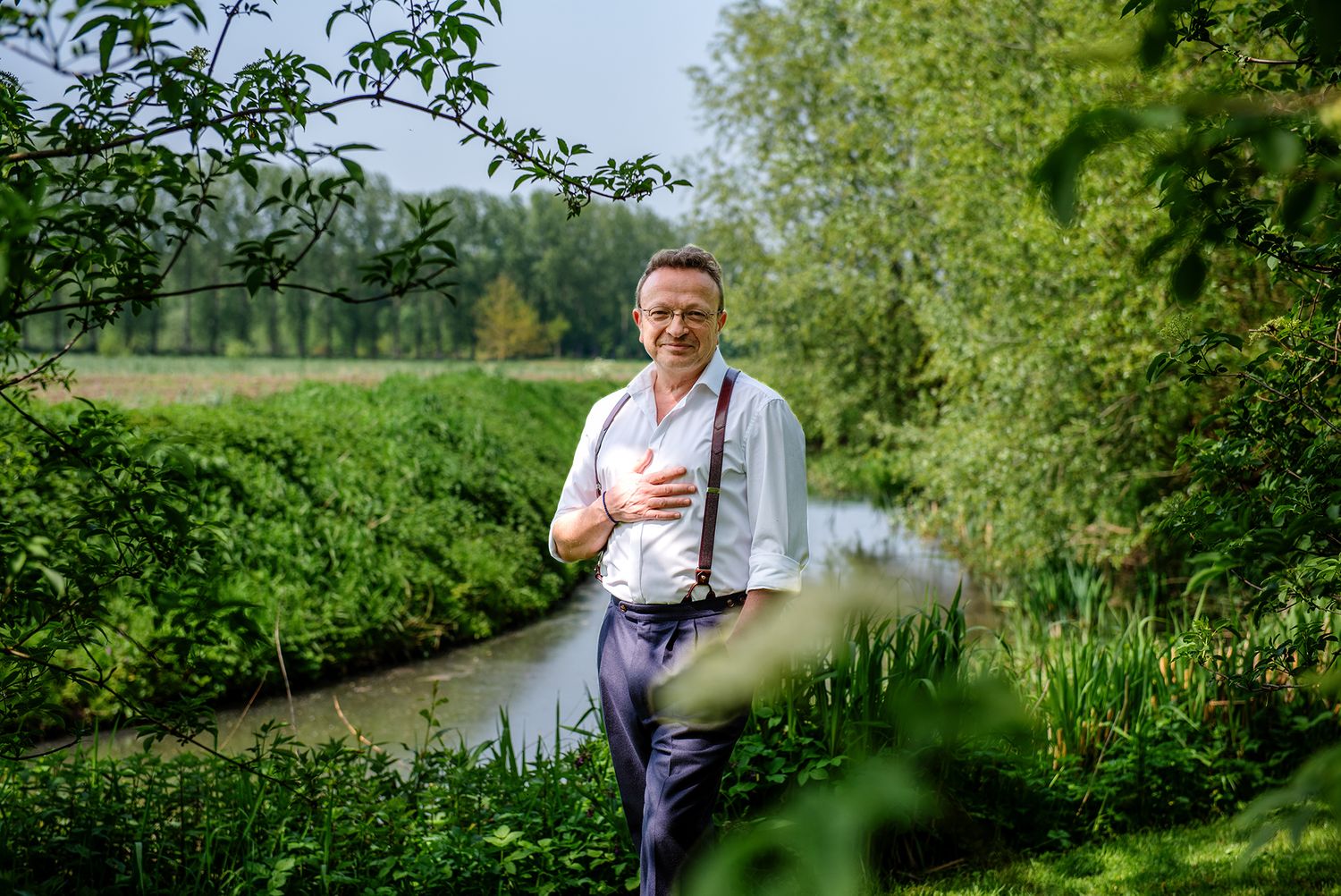 Middle-aged man in white shirt and suspenders smiling by a scenic green river landscape.