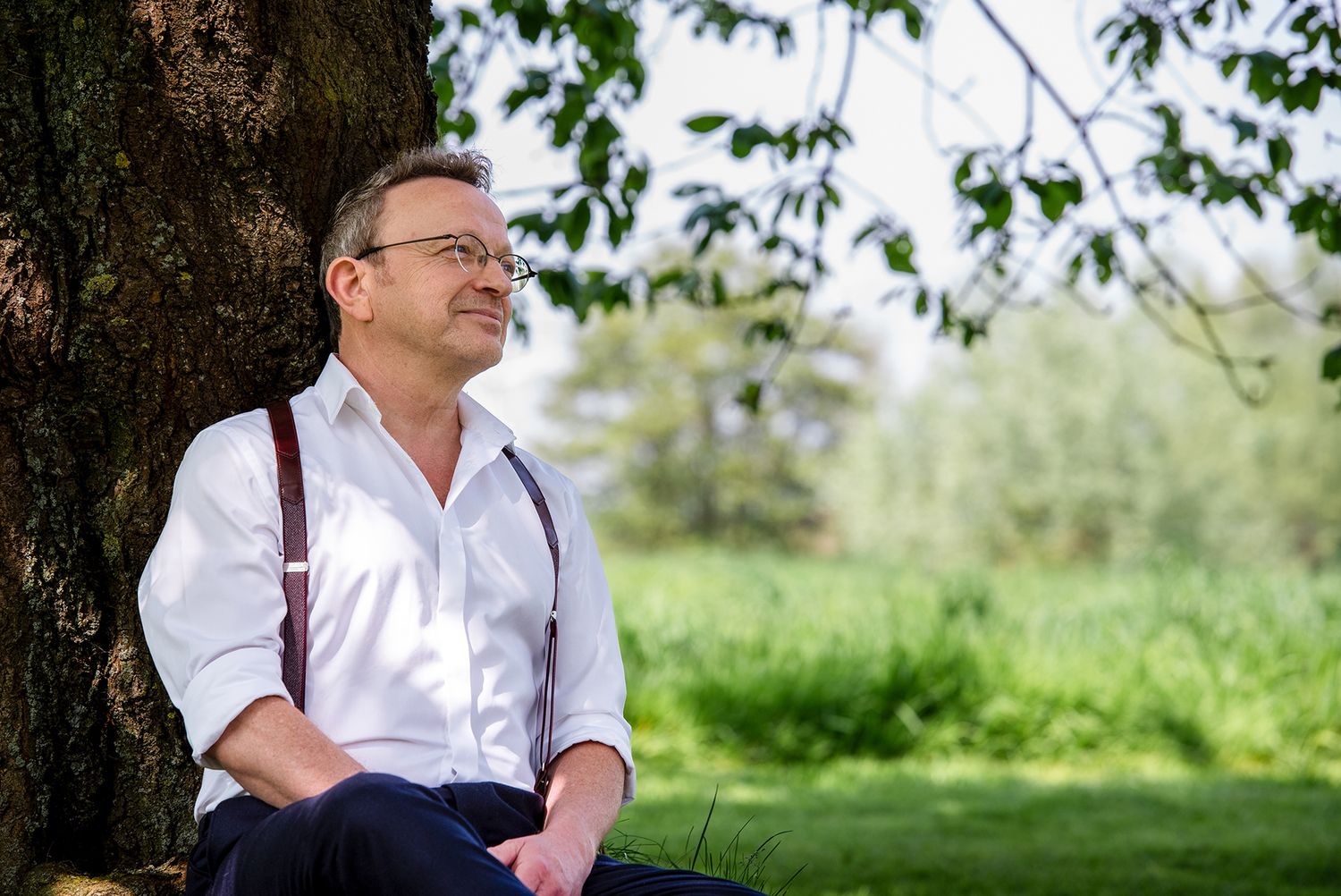 Thoughtful man in white shirt leaning against a tree trunk in a serene green outdoor setting.