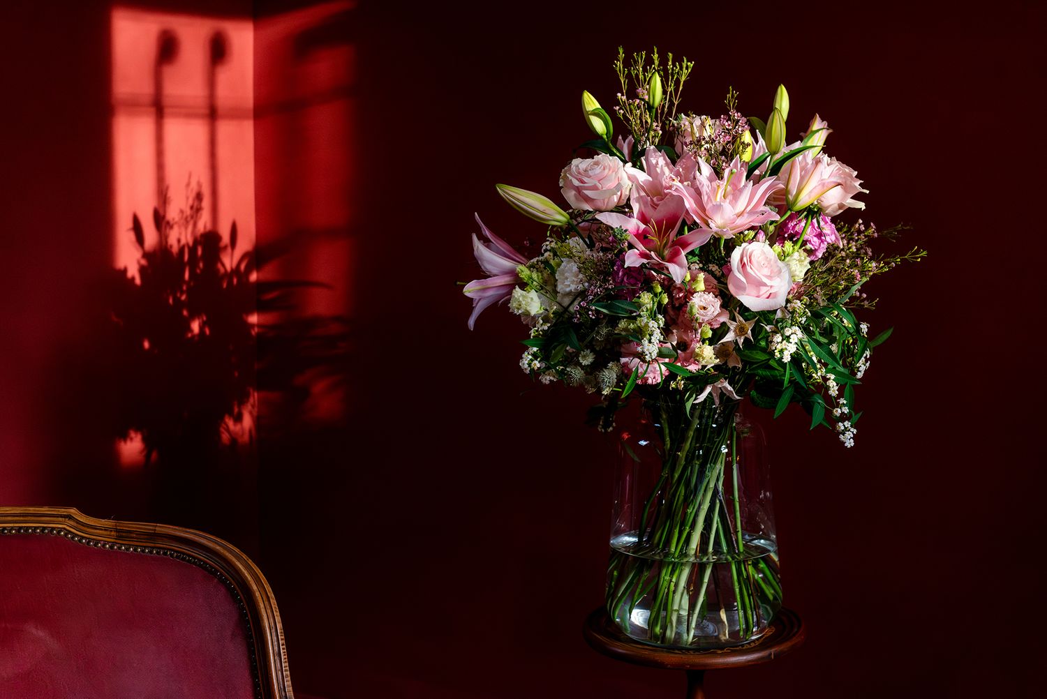 Lush pink bouquet of roses and lilies in glass vase against dramatic deep red wall with chair.