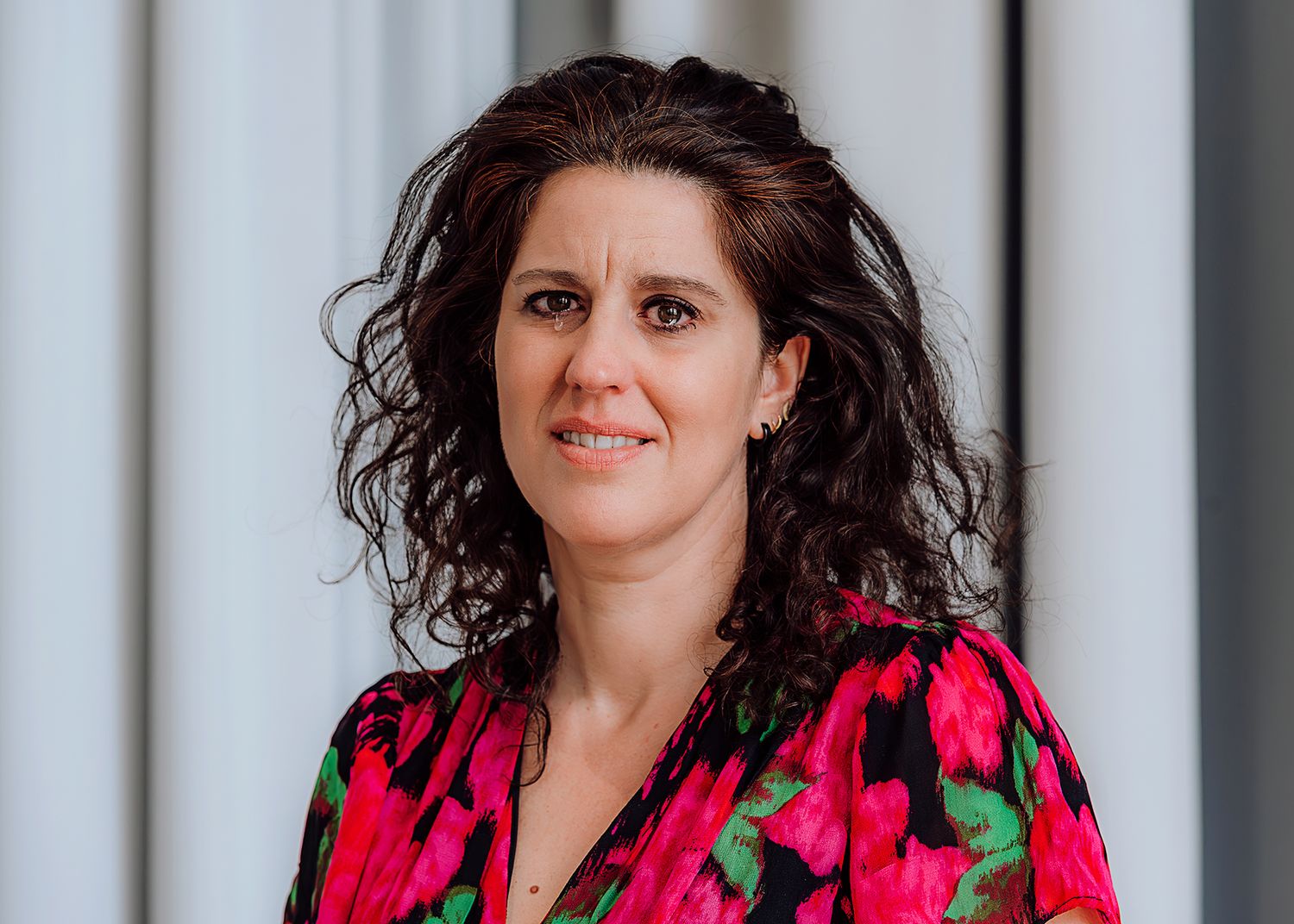 A woman with curly dark hair smiles warmly, wearing a vibrant floral blouse in a bright indoor setting.