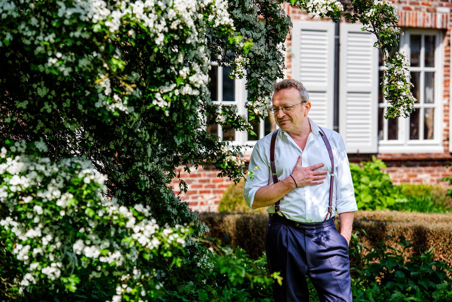 Man in white shirt and suspenders laughing beside white flowering shrubs near a brick building.