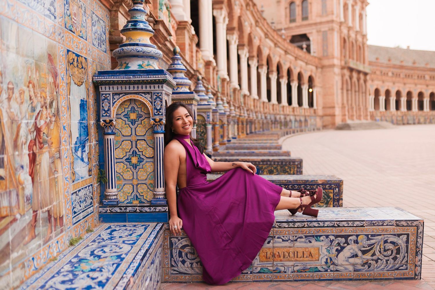 Woman in purple dress reclines on ornate tiled bench at Plaza de España, Seville, smiling at camera.