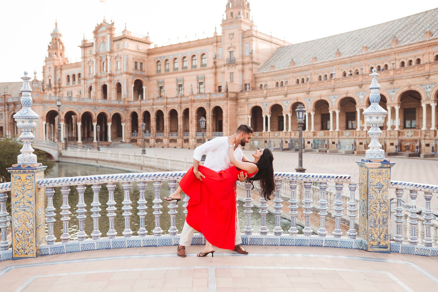 Couple dancing a dramatic dip in red dress on ornate tiled balcony at Plaza de España, Seville, with grand facade behind them.