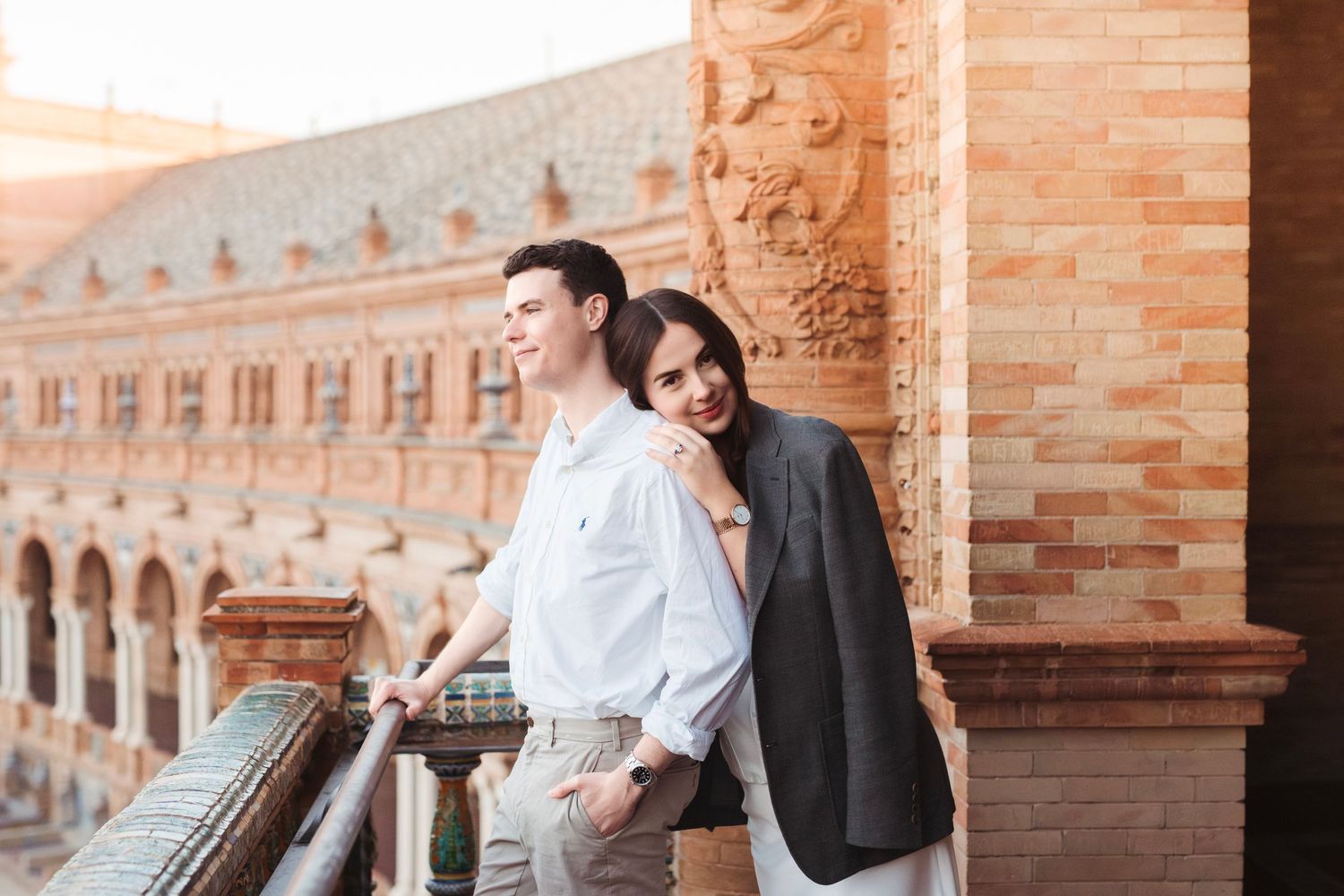 Couple leaning on a balcony railing at Plaza de España, Seville, with warm terracotta architecture behind.