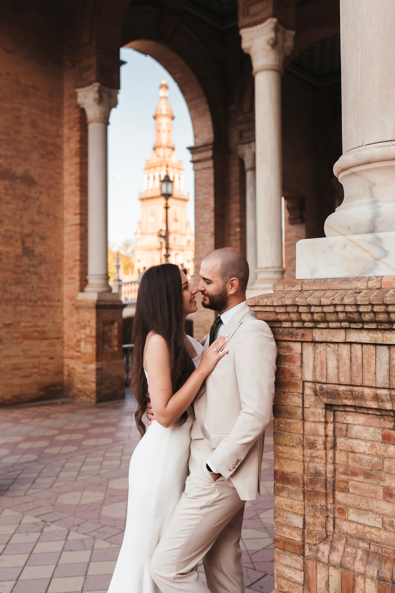 Wedding couple embracing under stone archway at Plaza de España, Seville, with tower visible in background.