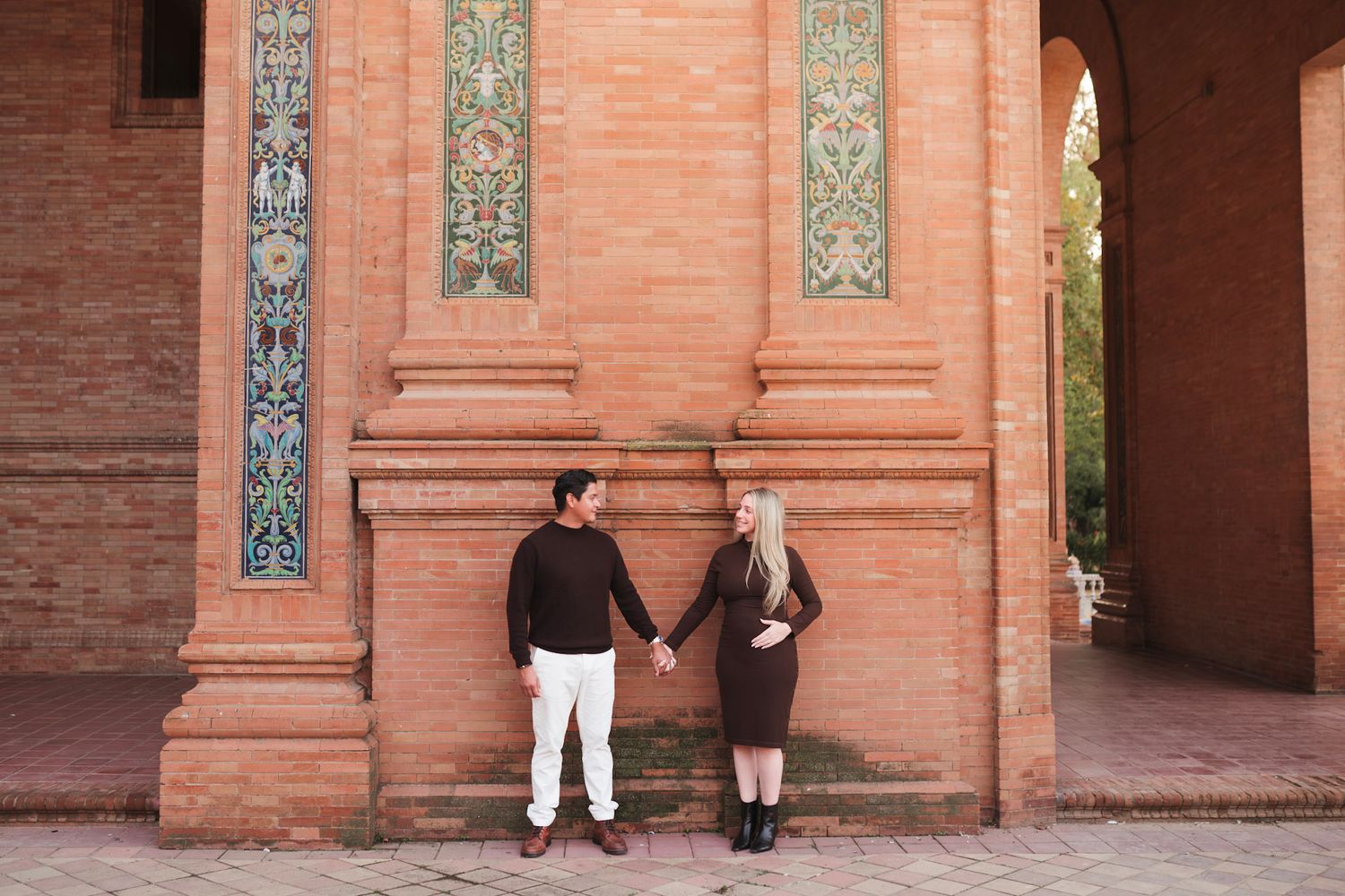 Expecting couple holding hands by ornate tiled wall at Plaza de España Seville.