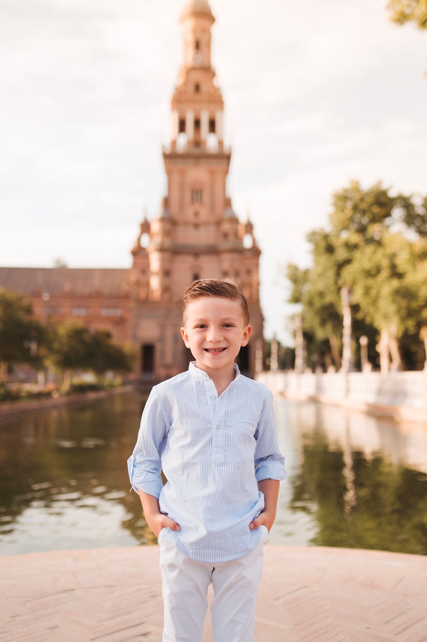 Smiling young boy in blue shirt standing near a canal with historic tower in background.