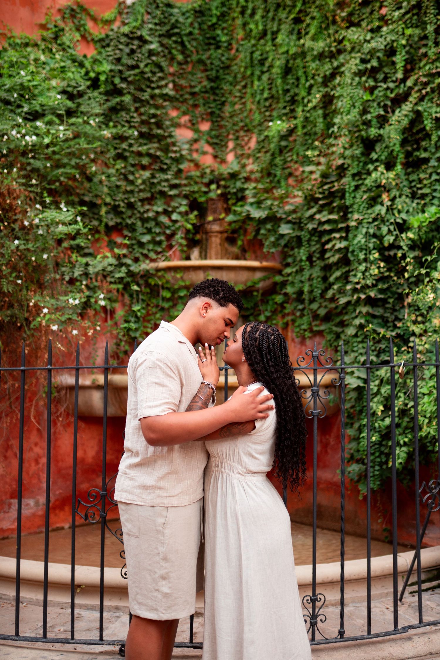 A couple shares a romantic moment near a stone fountain surrounded by lush green ivy on a red wall.