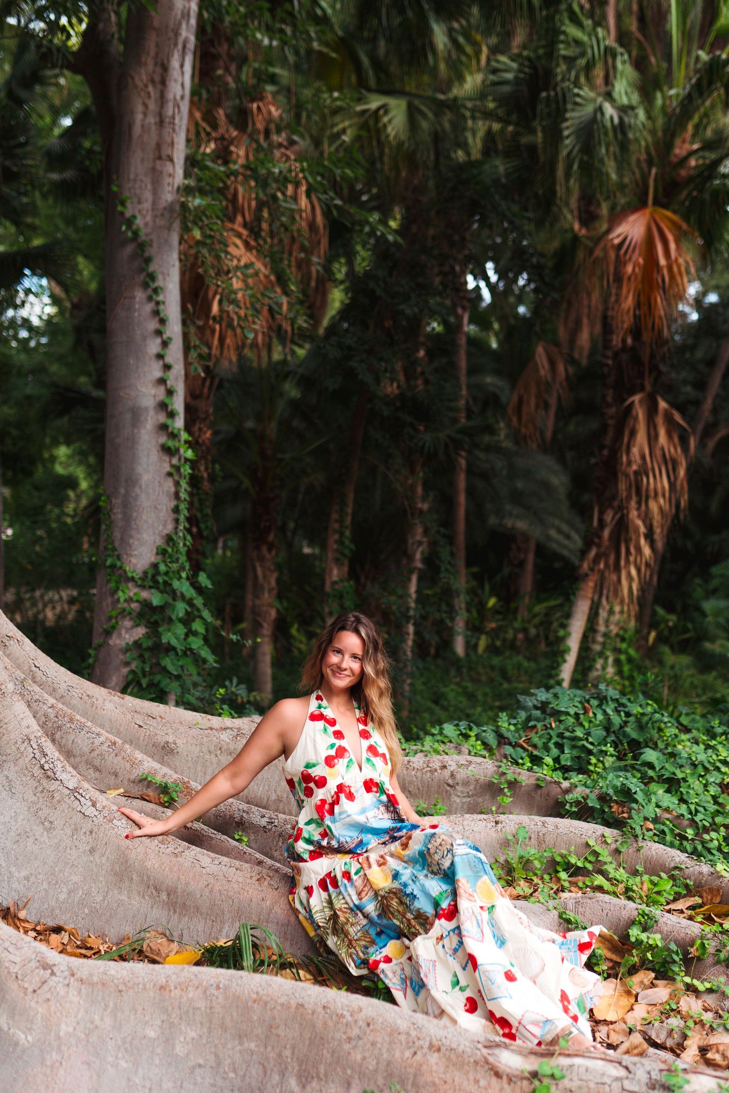 Woman in floral dress sitting on large tree roots surrounded by lush tropical palm trees in a garden.