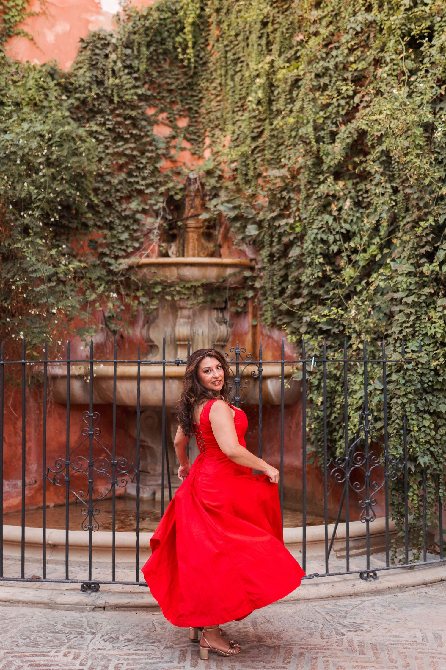 Woman in flowing red dress twirling near ornate iron gate with ivy-covered stone fountain backdrop.