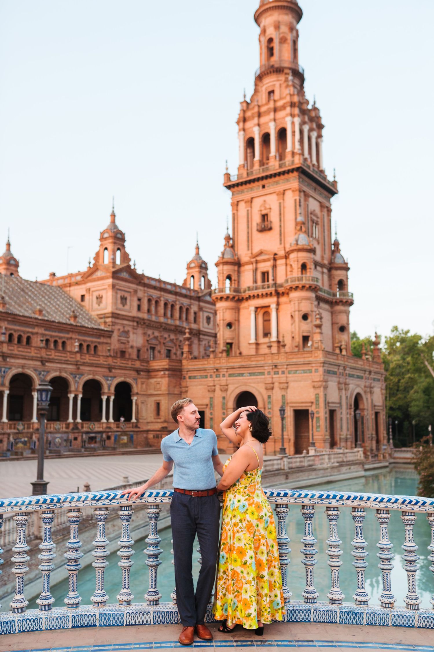 Couple embracing at Plaza de España in Seville, Spain, with ornate brick tower and tiled bridge in background.
