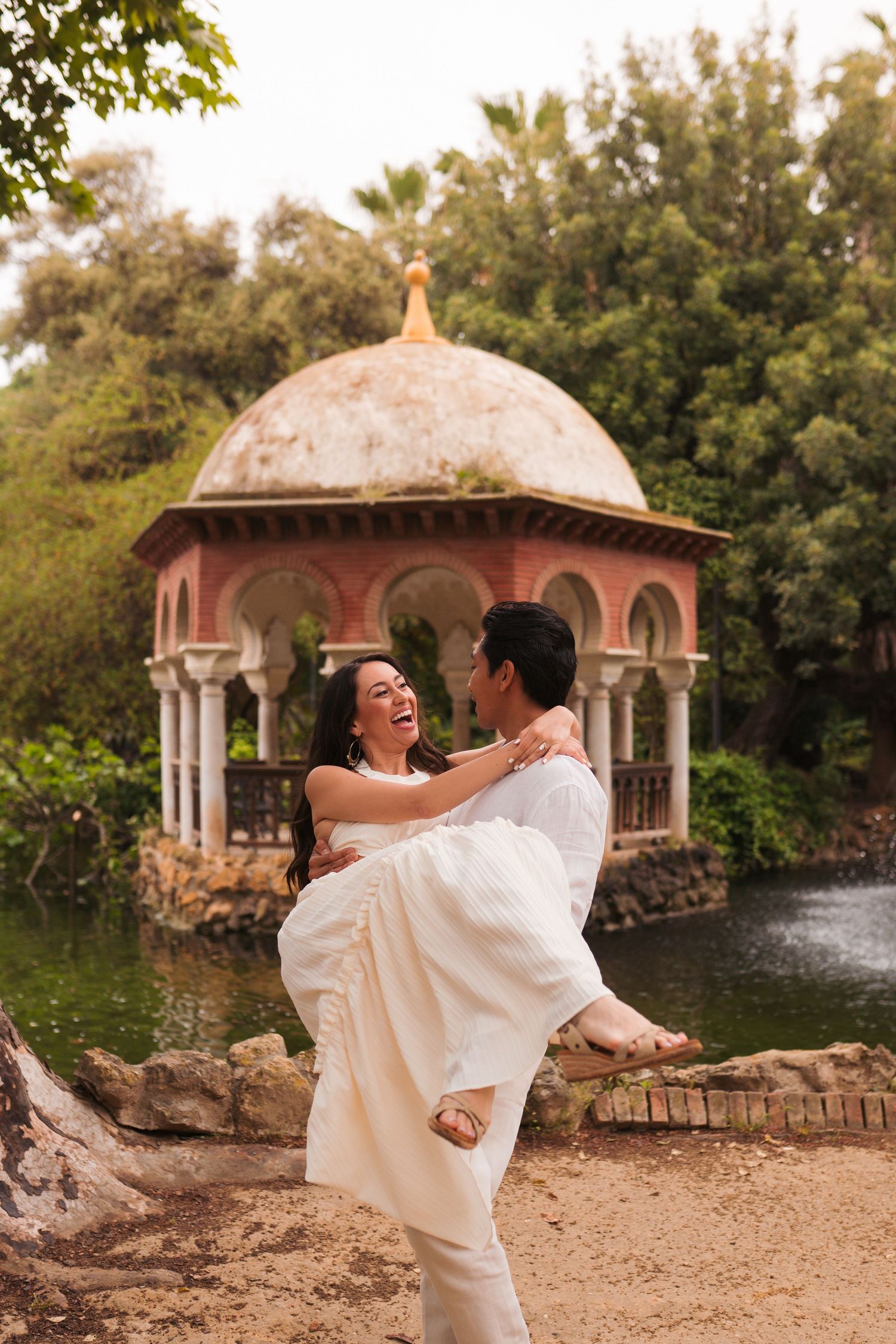 A joyful couple in white attire, man lifting woman, laughing near ornate gazebo and pond in lush garden.