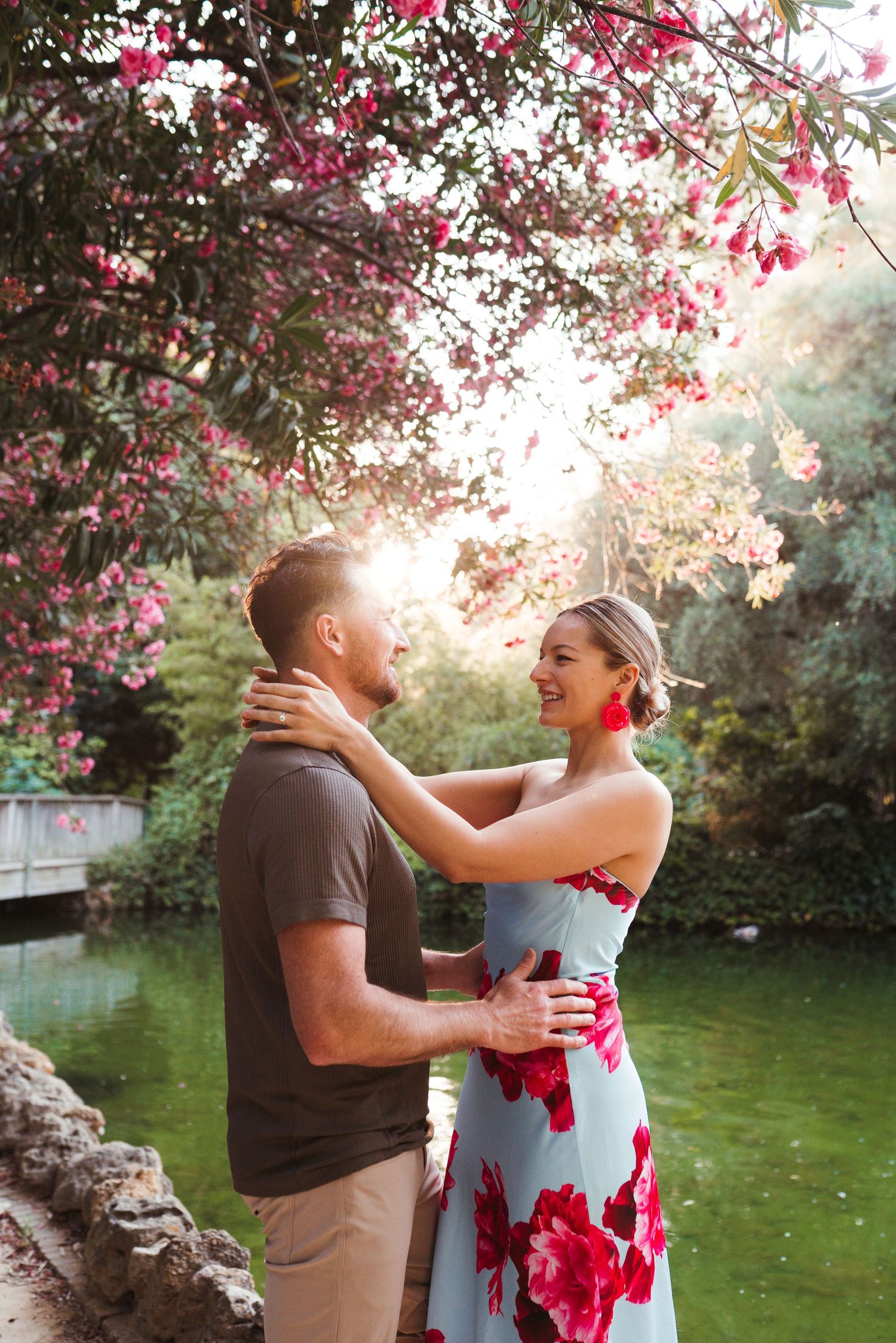 Couple embracing under pink flowering tree in a lush garden, woman in floral dress smiling at man in sunlit park.