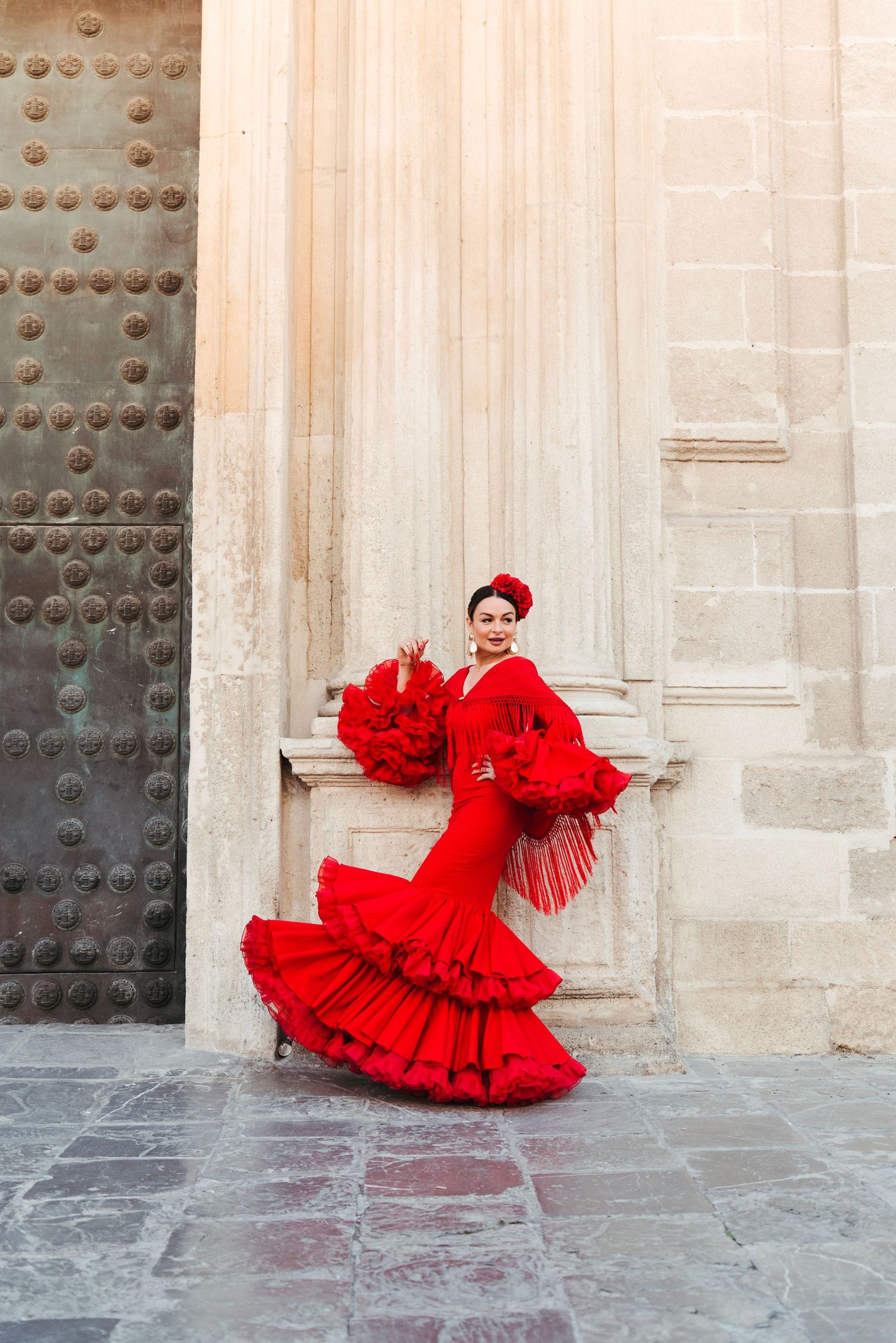 Woman in red flamenco dress poses against stone wall near ornate door in Spain.